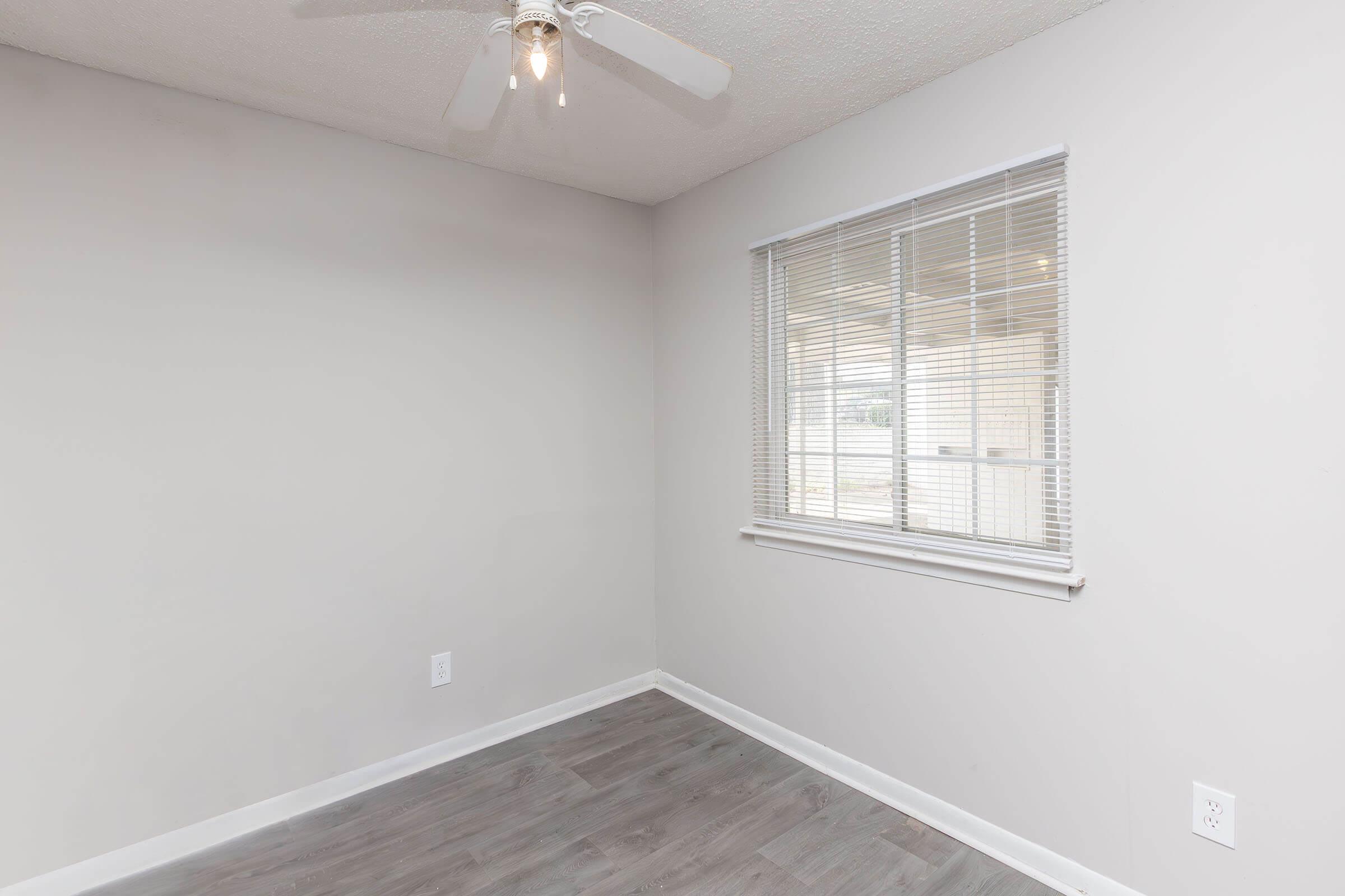 Empty room with light gray walls and a window featuring blinds. A ceiling fan with two lights is visible. The floor is a light wood-like laminate. The room has no furniture, creating a clean and open space.