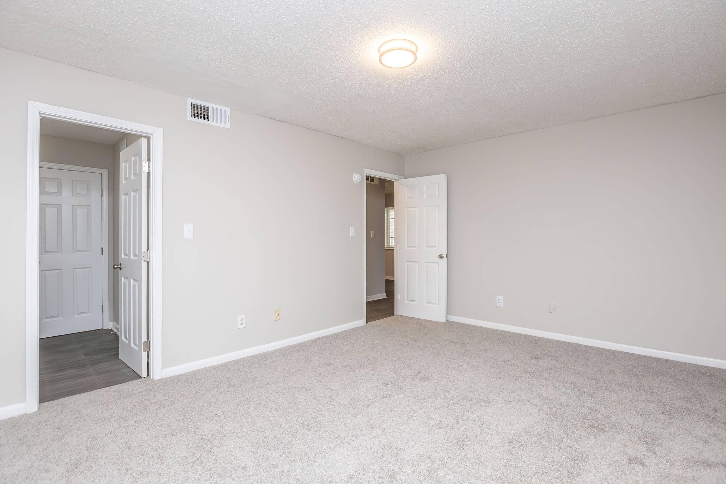 Spacious, empty room with light gray walls and carpeted flooring. Two white doors lead to adjacent rooms, and a ceiling light fixture provides illumination. The room is well-lit and has a neutral, clean aesthetic, making it ideal for various purposes.