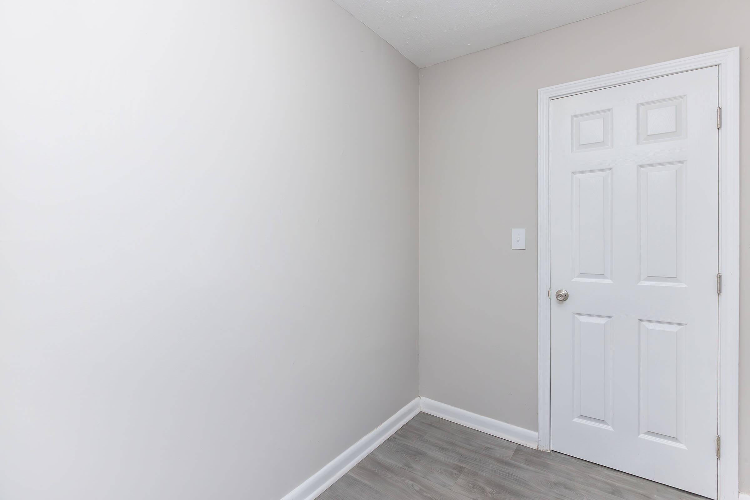 A minimalist interior corner of a room featuring light gray walls and a white door. The floor is covered with light-colored laminate. The overall design is simple and modern, evoking a clean and open space.
