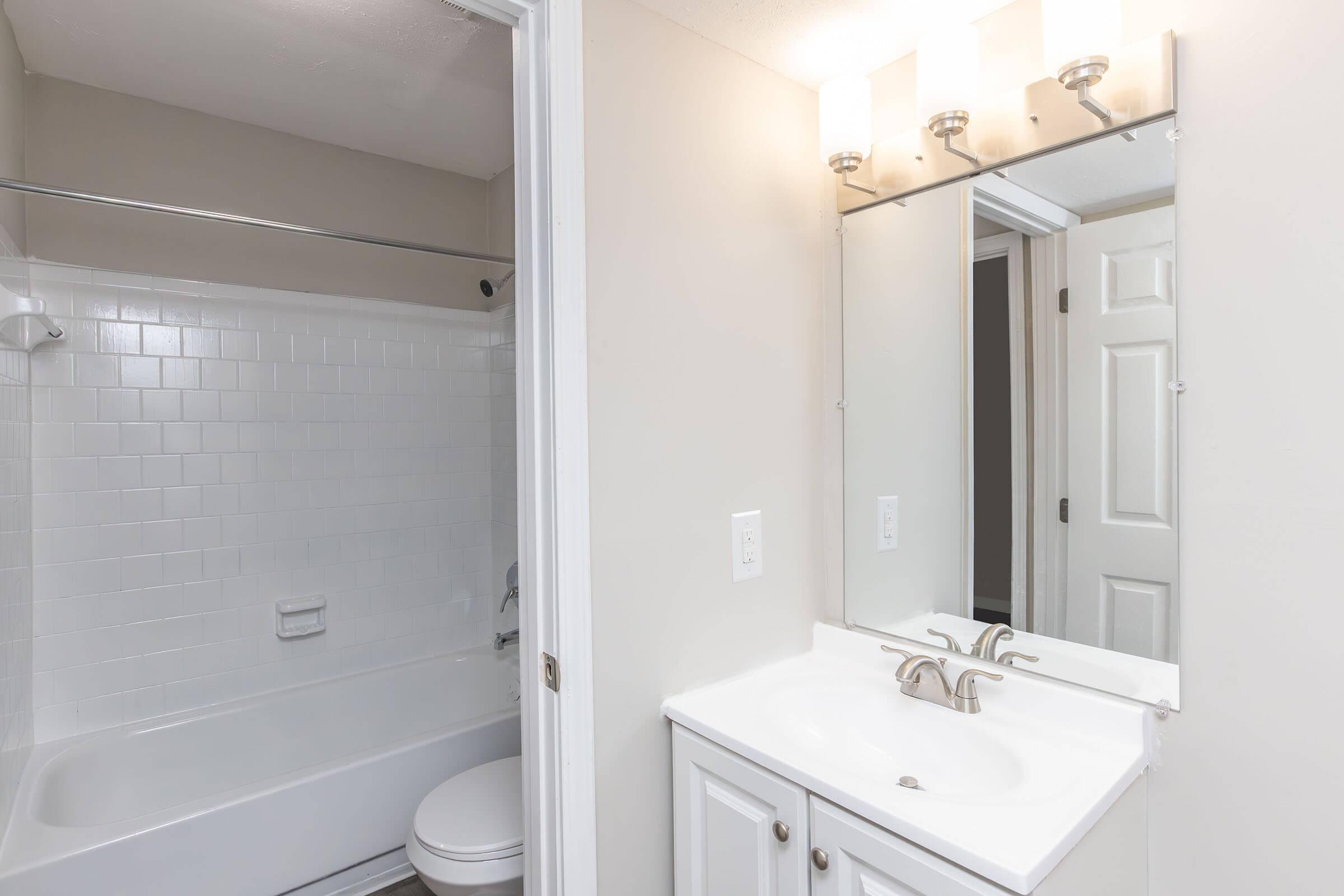 A well-lit bathroom featuring a white tub with a shower curtain, a white vanity with two sinks, and a large mirror above. The walls are painted in a light neutral color, and there is a door leading to another room visible in the background.