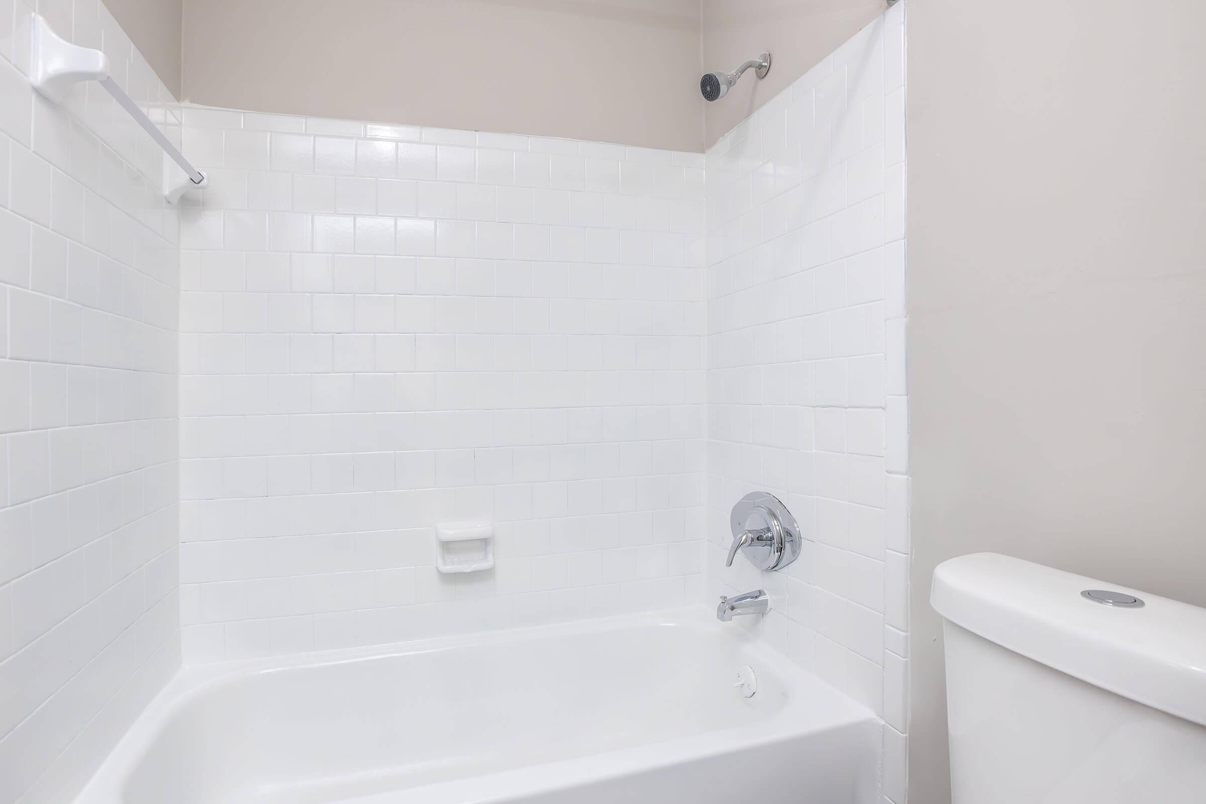 A clean bathroom featuring a white tiled shower area with a bathtub, a chrome showerhead, and a simple white toilet. The walls are painted a light color, creating a bright and modern look.