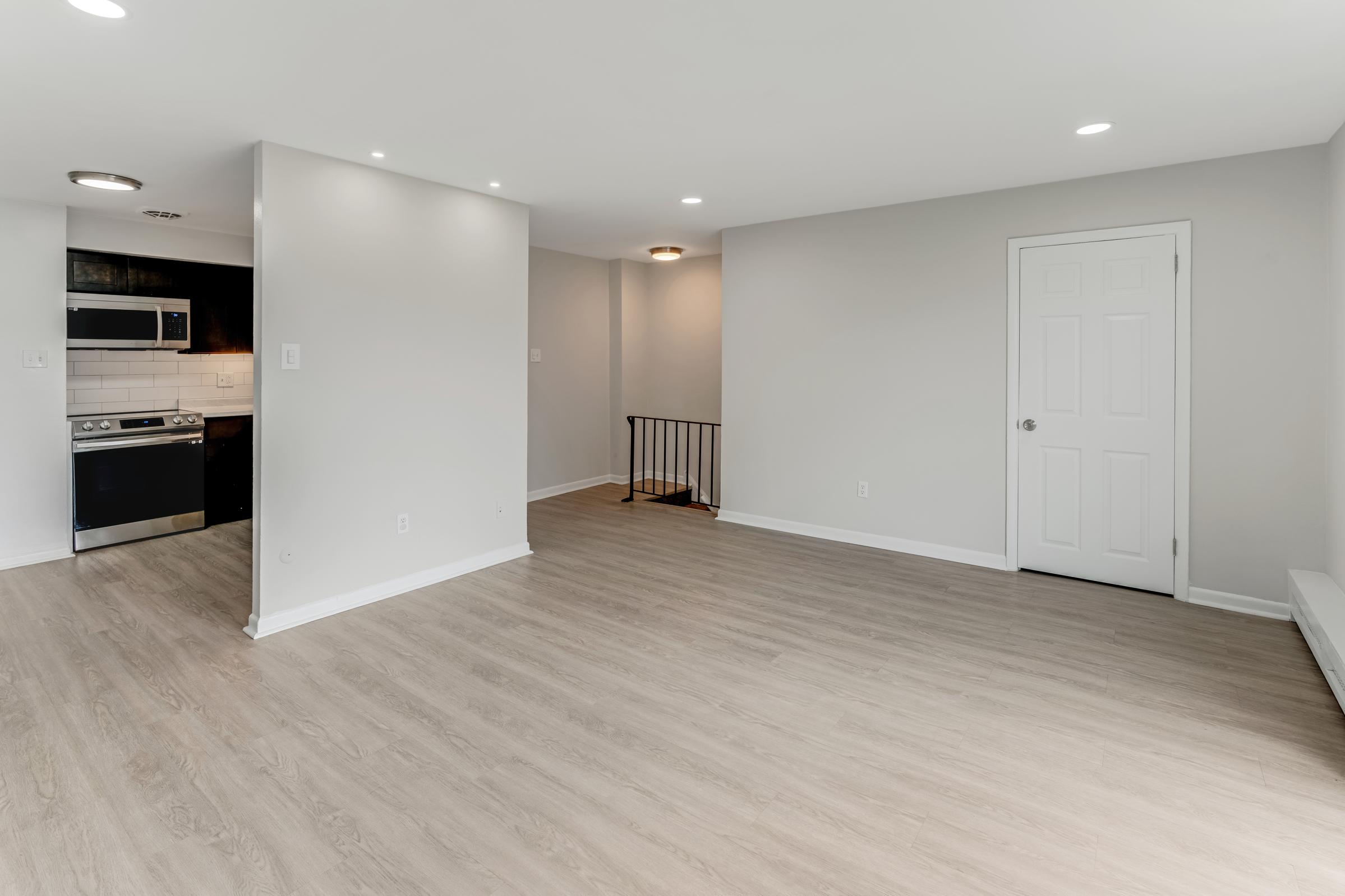 A spacious, well-lit living area with light wood flooring. The room features a doorway leading to a staircase, a modern kitchen visible in the background with stainless steel appliances, and neutral-colored walls that create an airy atmosphere.