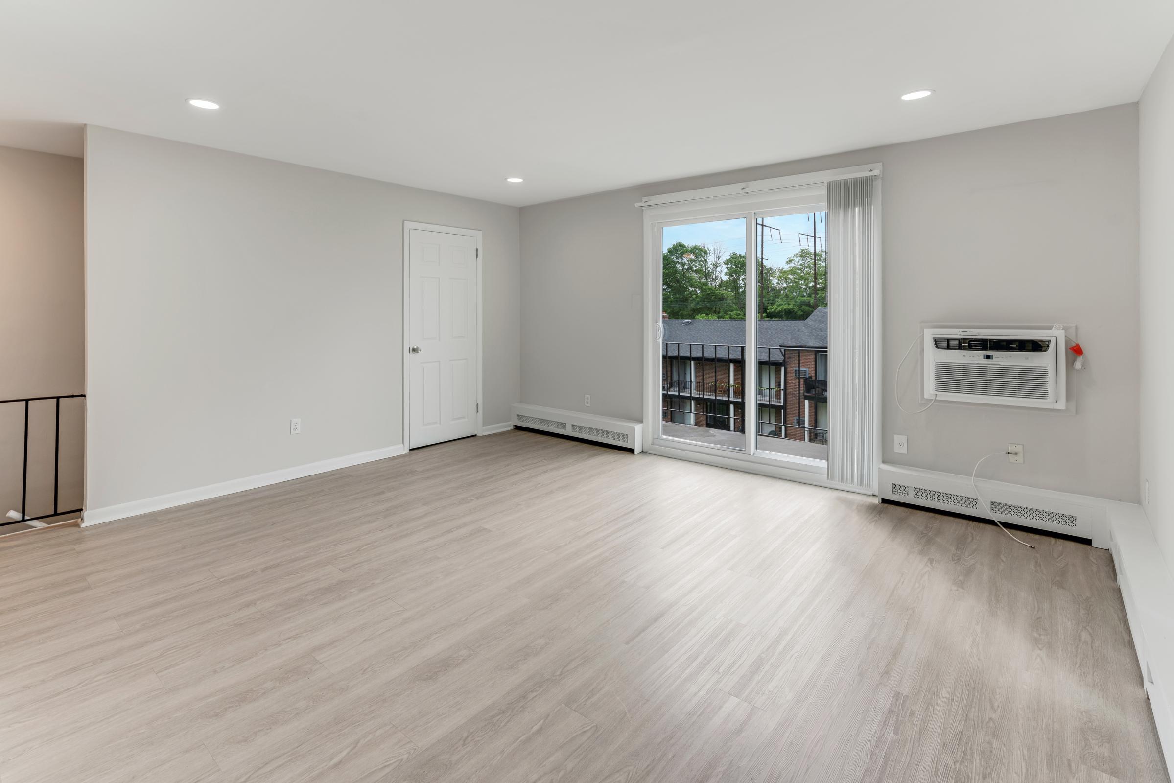 A spacious, empty room with light-colored walls and a light wood floor. There is a large window with sheer curtains allowing natural light, an air conditioning unit mounted on the wall, and a closed door leading to another room. The overall atmosphere is bright and modern, ideal for a living space.