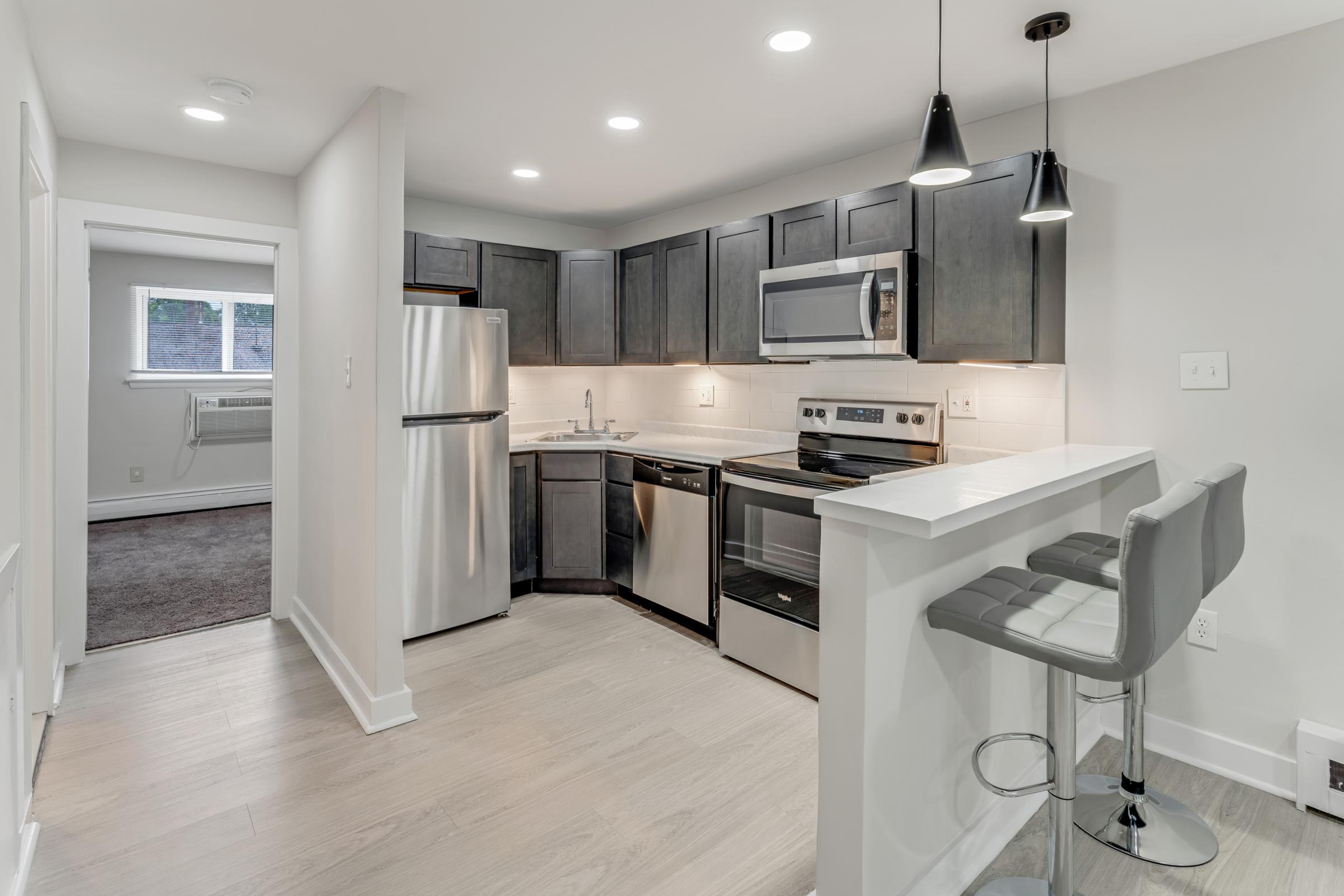 Modern kitchen interior featuring dark cabinetry, stainless steel appliances, and a bar area with two high stools. Soft lighting from pendant fixtures and recessed lights creates a cozy atmosphere. A window offers natural light, and light-colored flooring enhances the space.