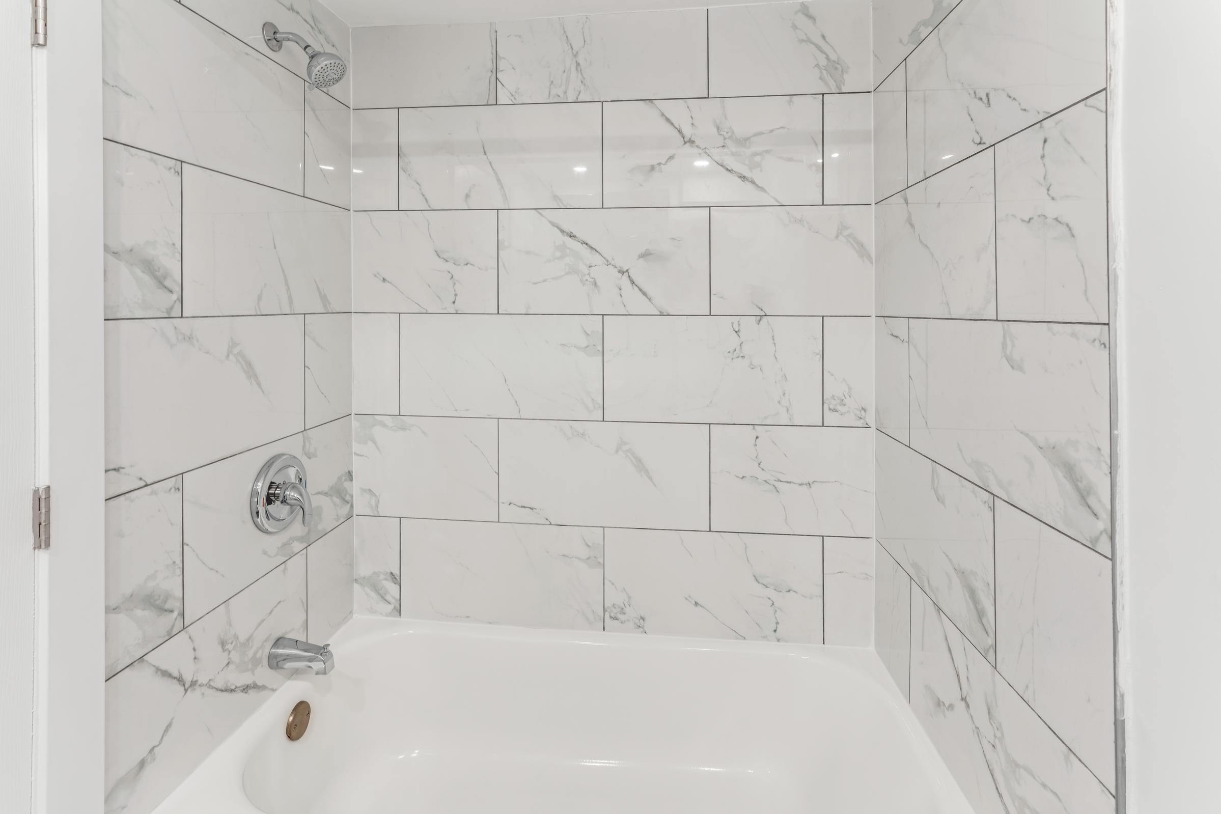 A modern shower with a white bathtub and marble-patterned tiles on the walls. The tiles feature a light gray and white veining design, and there is a chrome showerhead installed above. The overall aesthetic is sleek and clean.