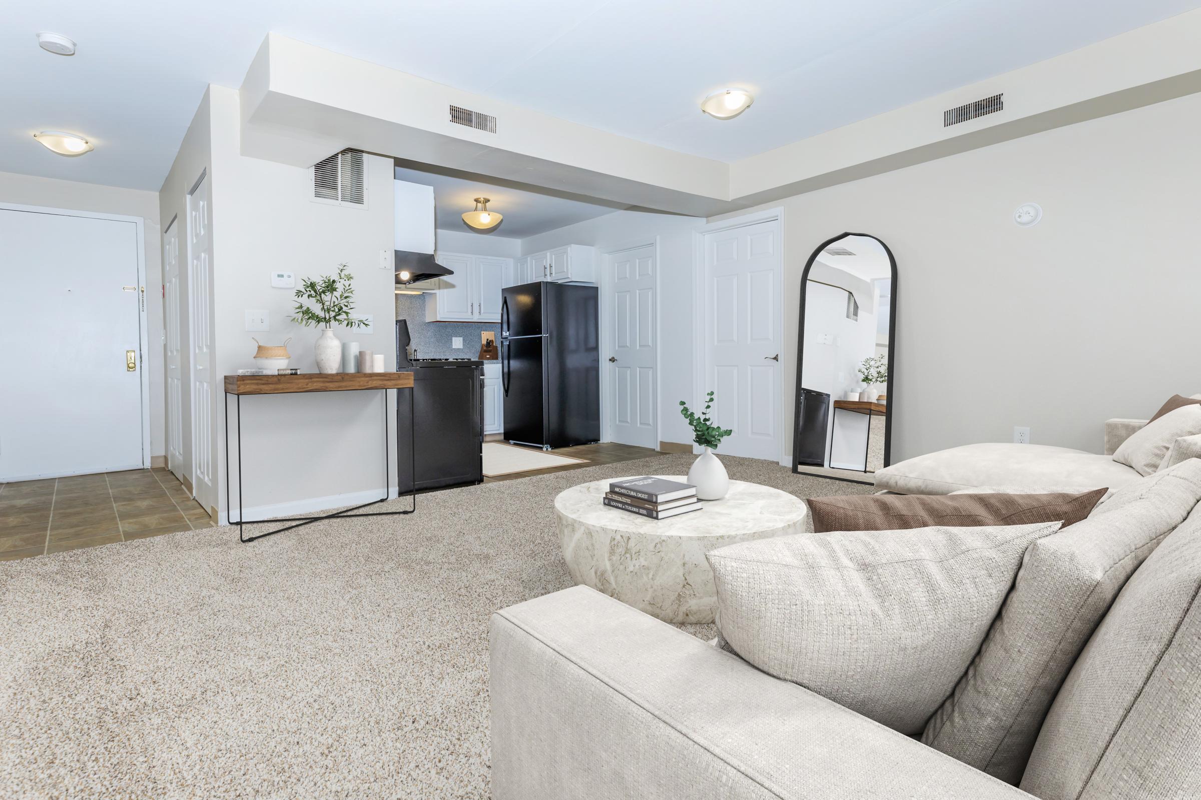 A modern living room featuring a light-colored sectional sofa and a round coffee table. There is a large mirror reflecting the space. The kitchen area is visible in the background, with dark cabinetry and appliances. Soft lighting creates a warm and inviting atmosphere, complemented by neutral wall colors and carpet.