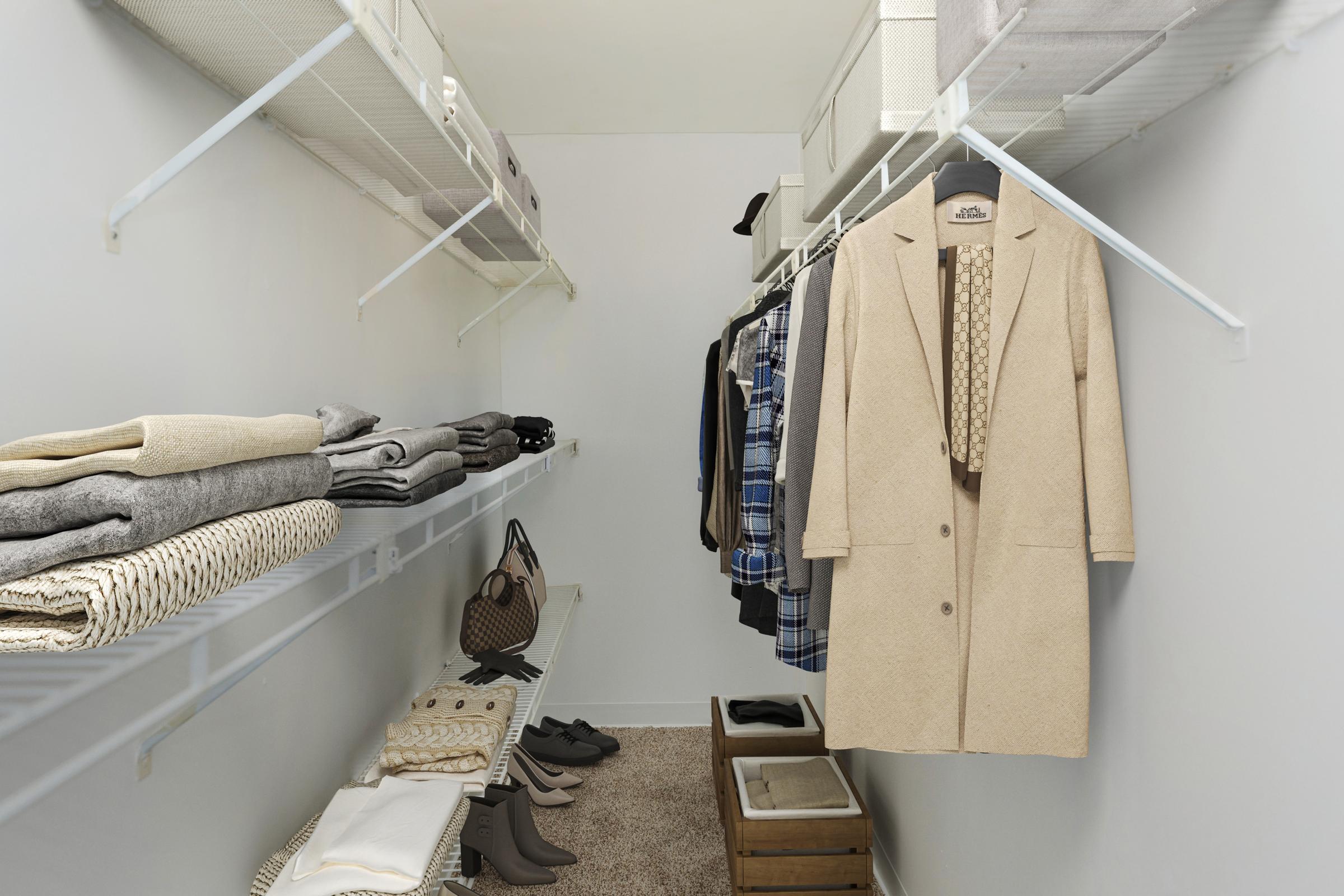 A neatly organized closet with shelves displaying folded sweaters, a light beige coat hanging, and a variety of clothing items including plaid and patterned garments. There are shoes on the floor and a few storage bins, all set against a clean, light-colored wall.