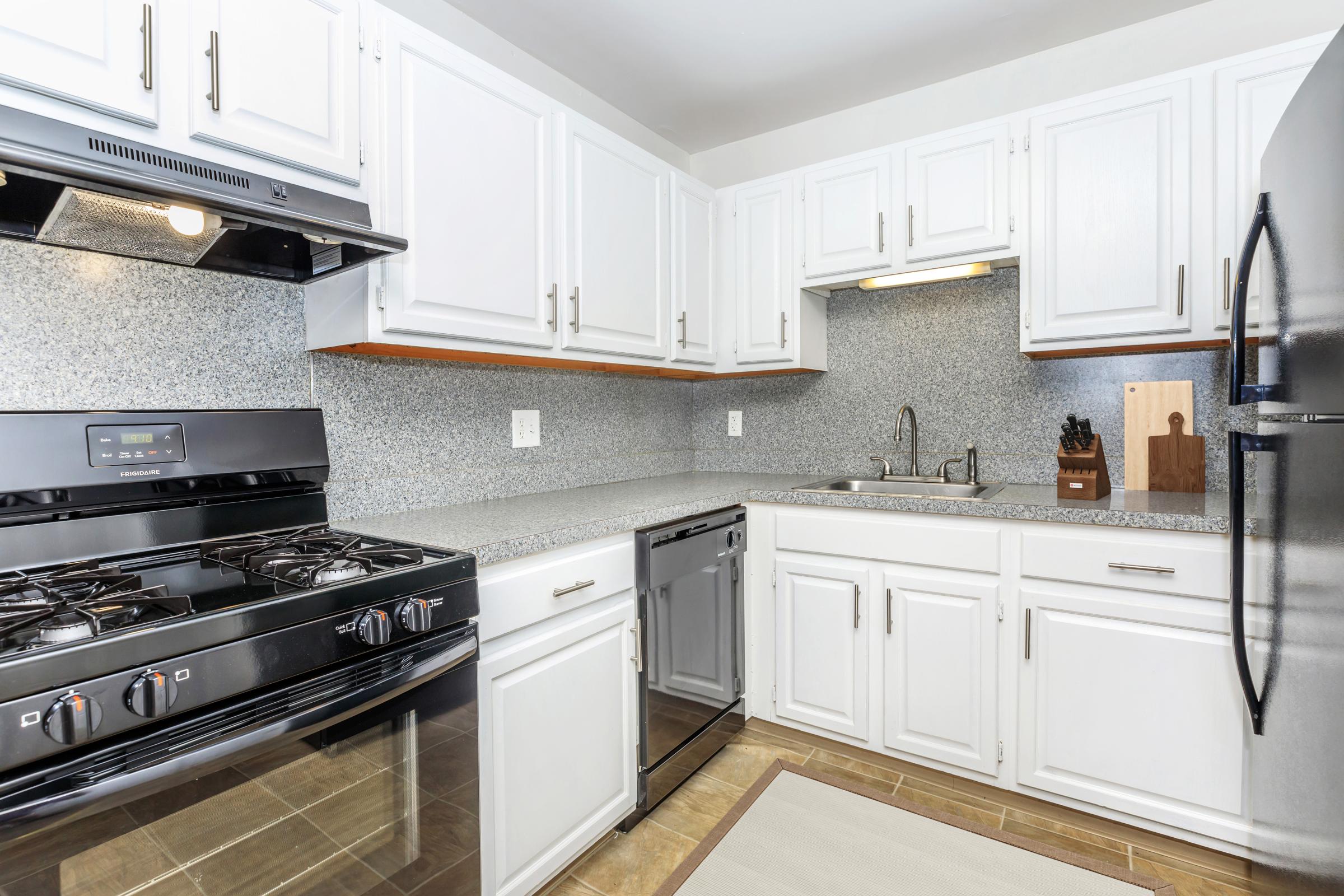 Modern kitchen featuring white cabinetry, stainless steel appliances, and a gray speckled countertop. Includes a gas stove, built-in dishwasher, and a kitchen sink with a faucet. The space is well-lit, showcasing a clean and organized layout with a neutral color scheme. A cutting board is visible on the countertop.