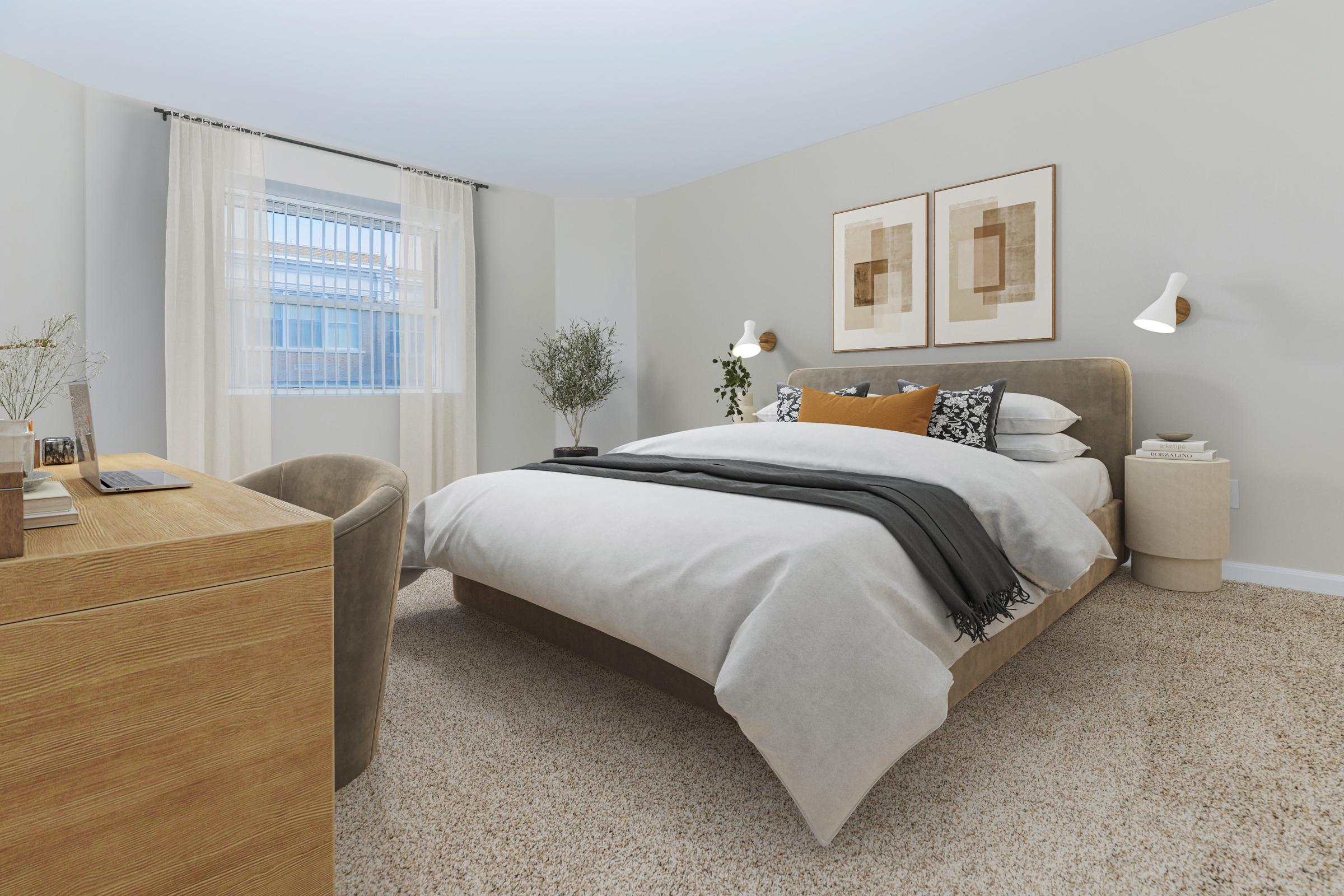 A cozy bedroom featuring a large bed with neutral bedding, a decorative throw, and pillows. There are two framed artworks on the wall, a small plant beside the window, and a wooden desk with a chair. Soft curtains let in natural light, creating a warm and inviting atmosphere. The carpet adds a touch of comfort.