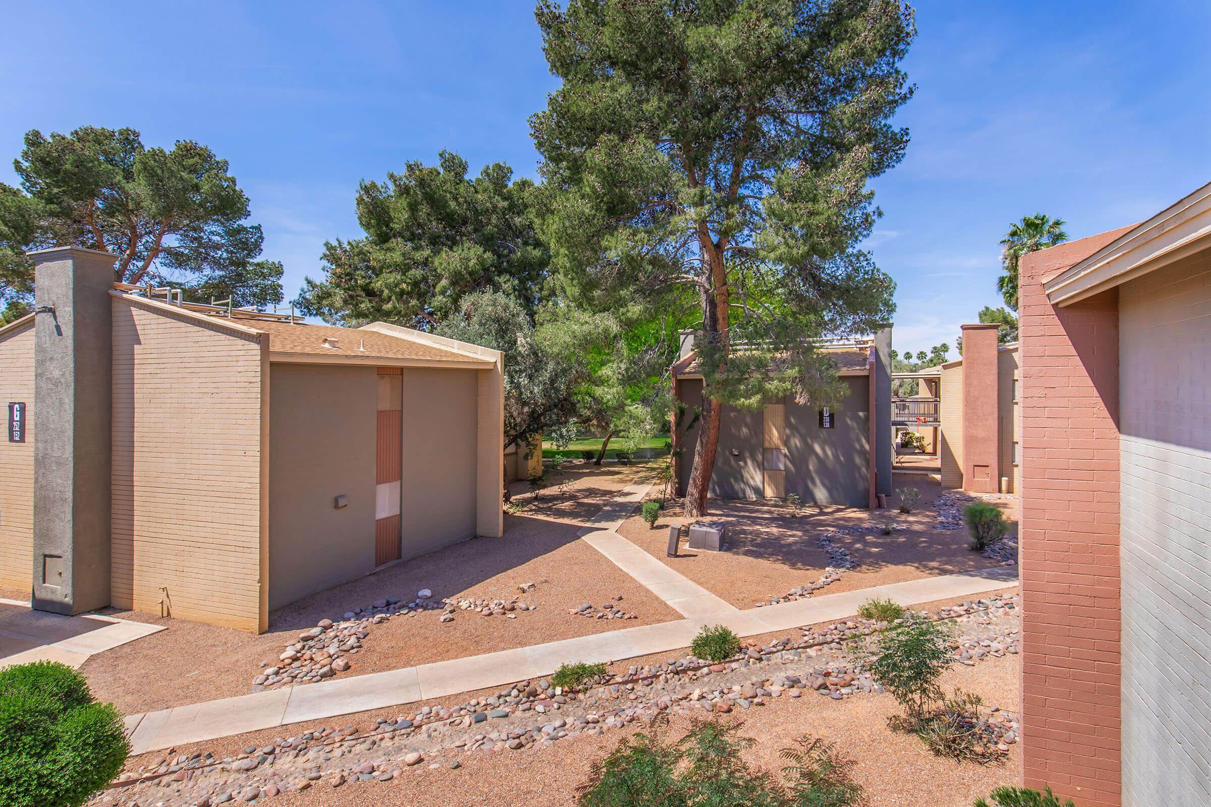 A view of a residential area featuring beige and brown buildings surrounded by gravel pathways, bushes, and trees. The sky is clear with a few clouds, and the landscape appears well-maintained and inviting, suggesting a peaceful atmosphere.