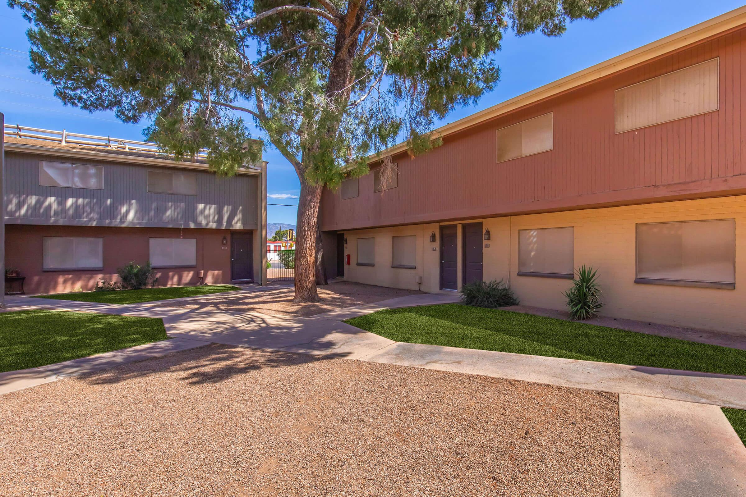 Two apartment buildings in a courtyard setting, surrounded by grass and gravel paths. A large tree provides shade. One building has a light-colored facade, while the other is a darker tone. The scene is bright and sunny, with a clear blue sky in the background.