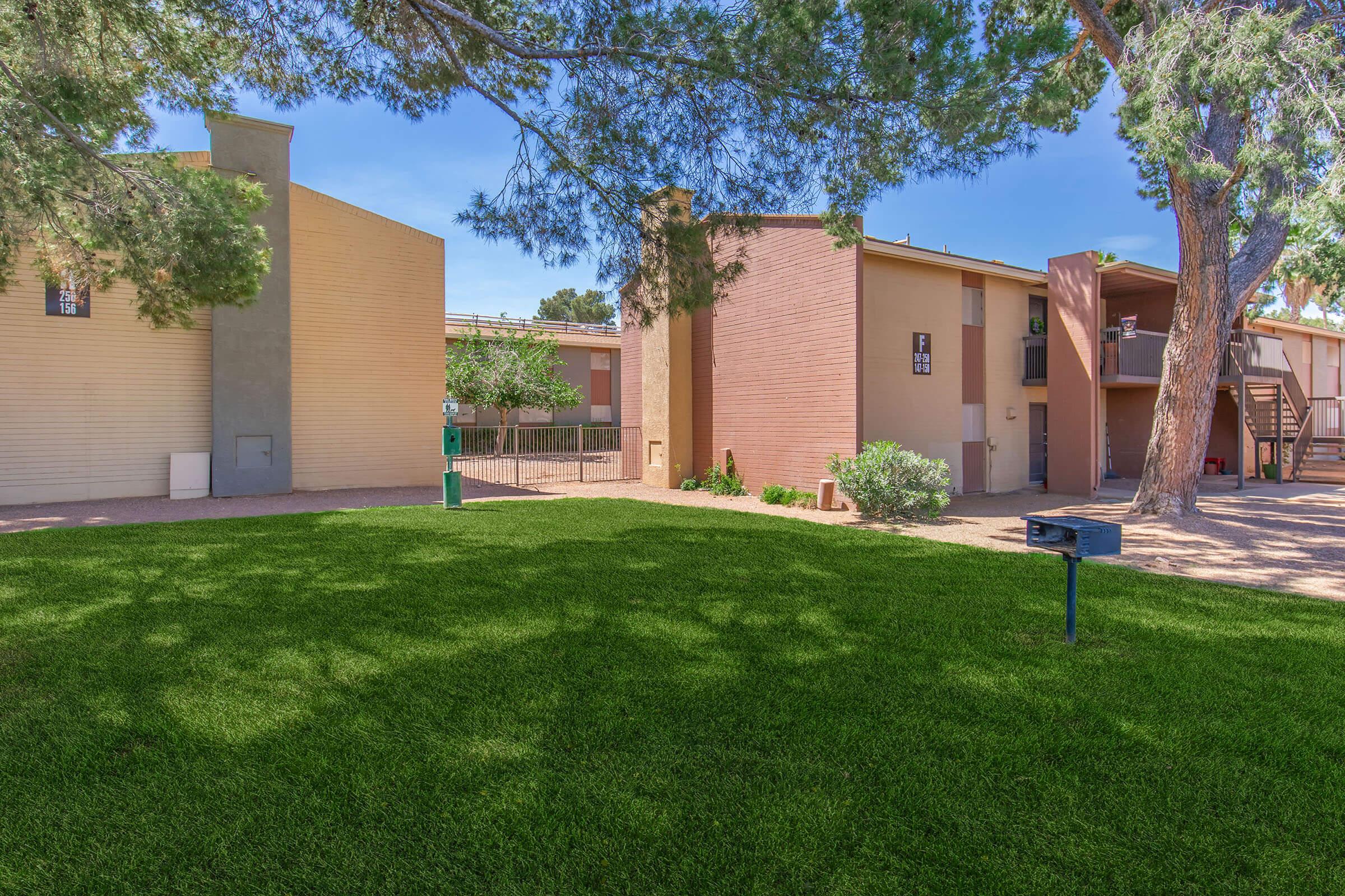 Two multi-unit residential buildings with a landscaped grassy area in front. The buildings have various shades of beige and brown exteriors, surrounded by trees. In the foreground, there is a mailbox on a post. The sky is clear and blue, creating a bright, inviting atmosphere.