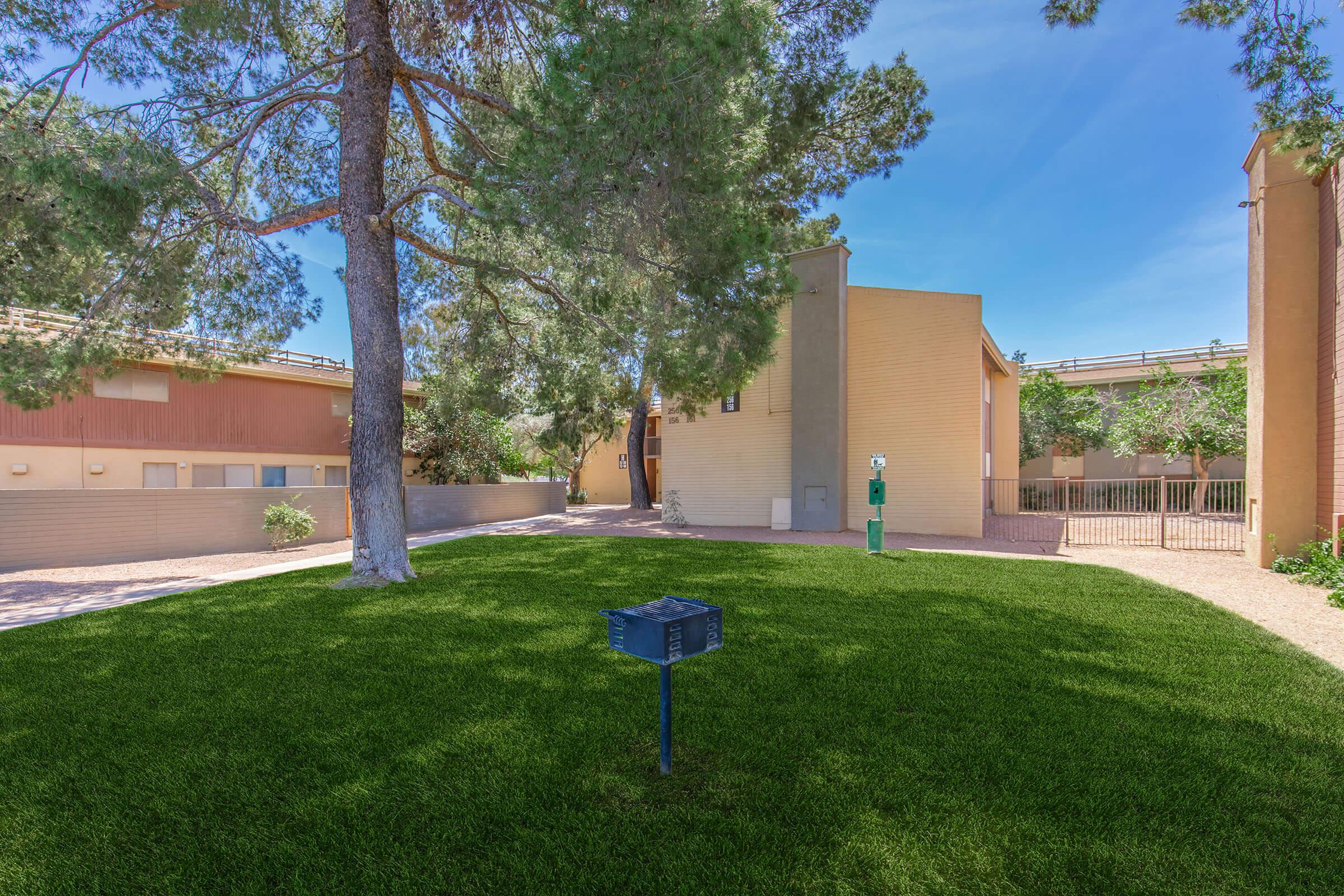 A well-maintained green lawn surrounded by trees and buildings. The area features a small black mailbox on a post, outdoor lighting, and a fence in the background, with a clear blue sky above. Ideal for outdoor activities and relaxation.