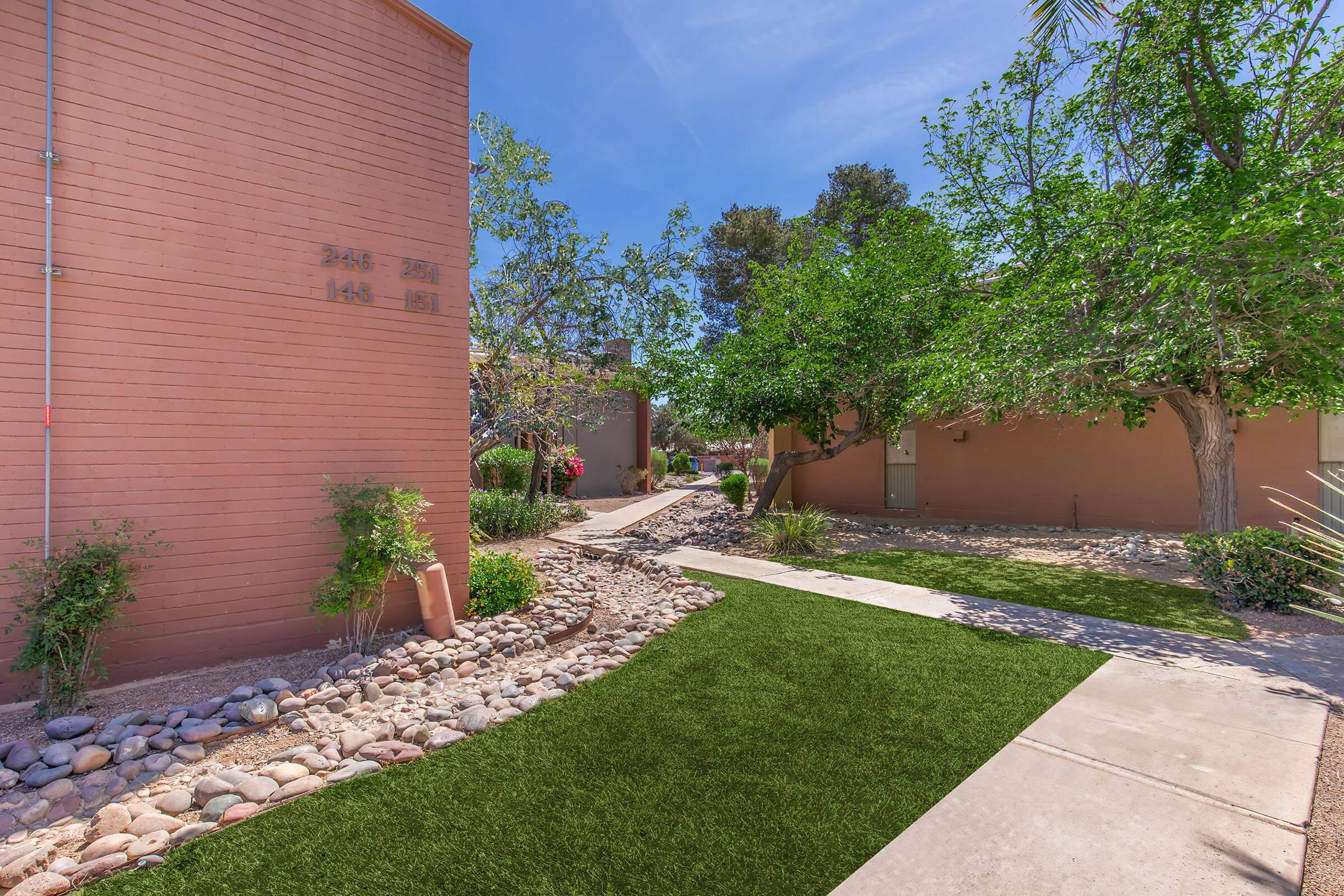 A landscaped pathway featuring artificial grass and pebbled borders, surrounded by lush greenery and trees. The pathway leads through a residential area with textured brick walls and sunlight casting shadows on the ground. Bright blue sky overhead enhances the tranquil atmosphere.