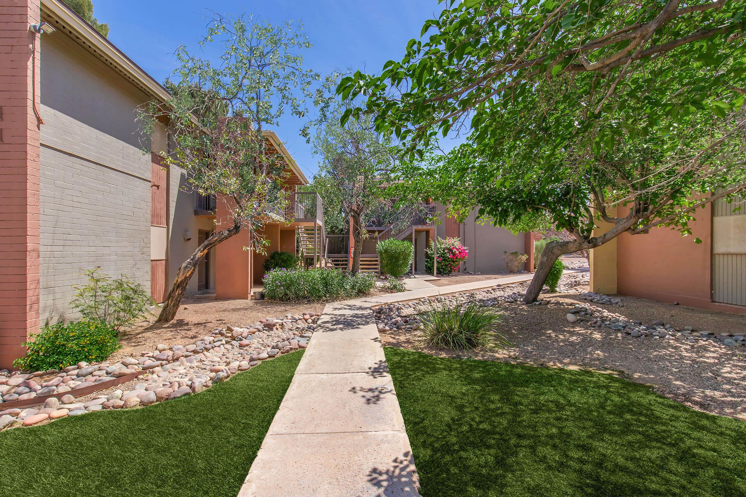 A landscaped pathway winding between two residential buildings, lined with grass and decorative rocks. Lush green trees and colorful plants add to the vibrant, tranquil atmosphere of the outdoor space. The clear blue sky enhances the bright and inviting environment.