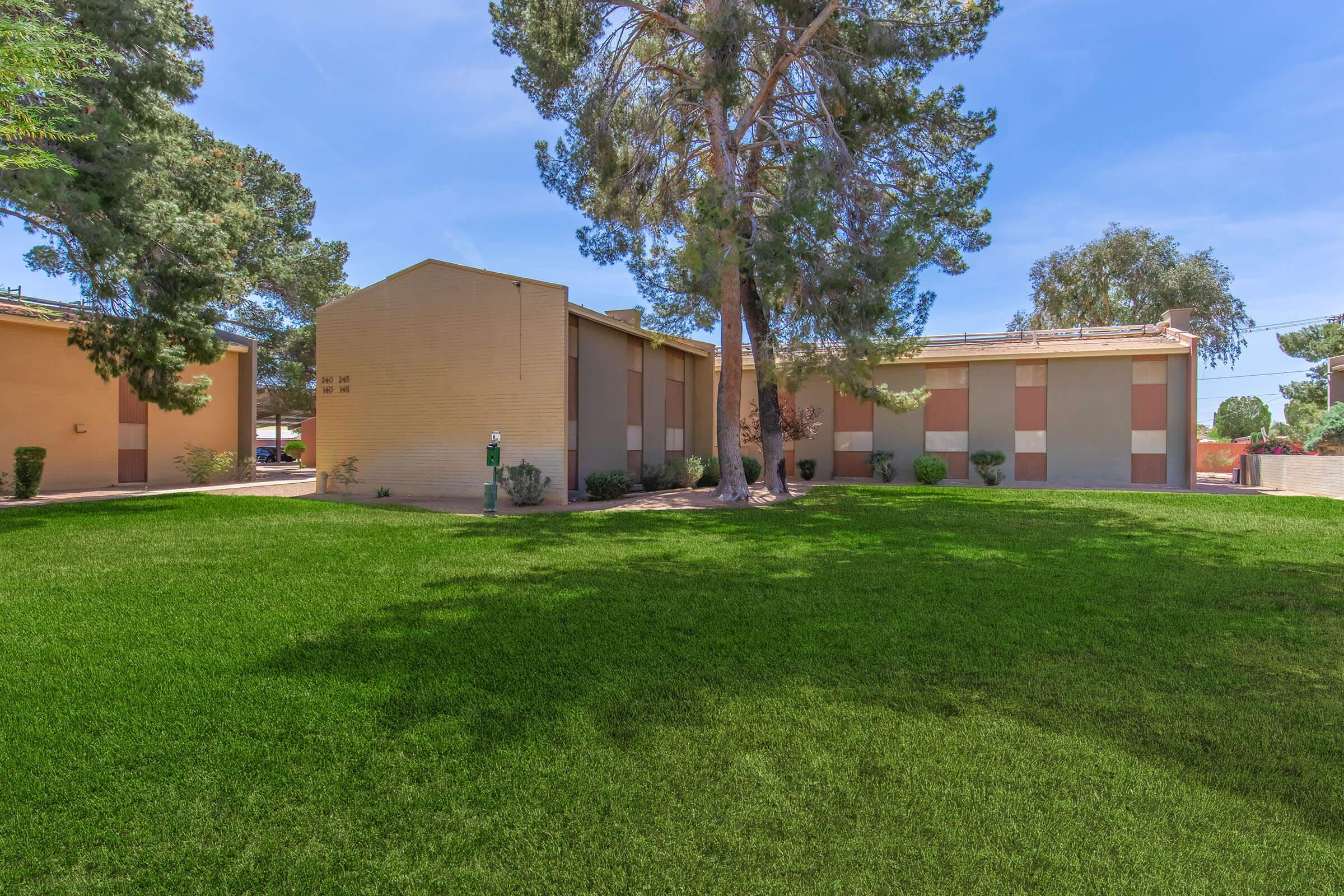 A view of a well-maintained building surrounded by lush green grass and several trees. The architecture features a mix of beige and pastel colors, with landscaped areas and a clear blue sky in the background, creating a peaceful outdoor setting.
