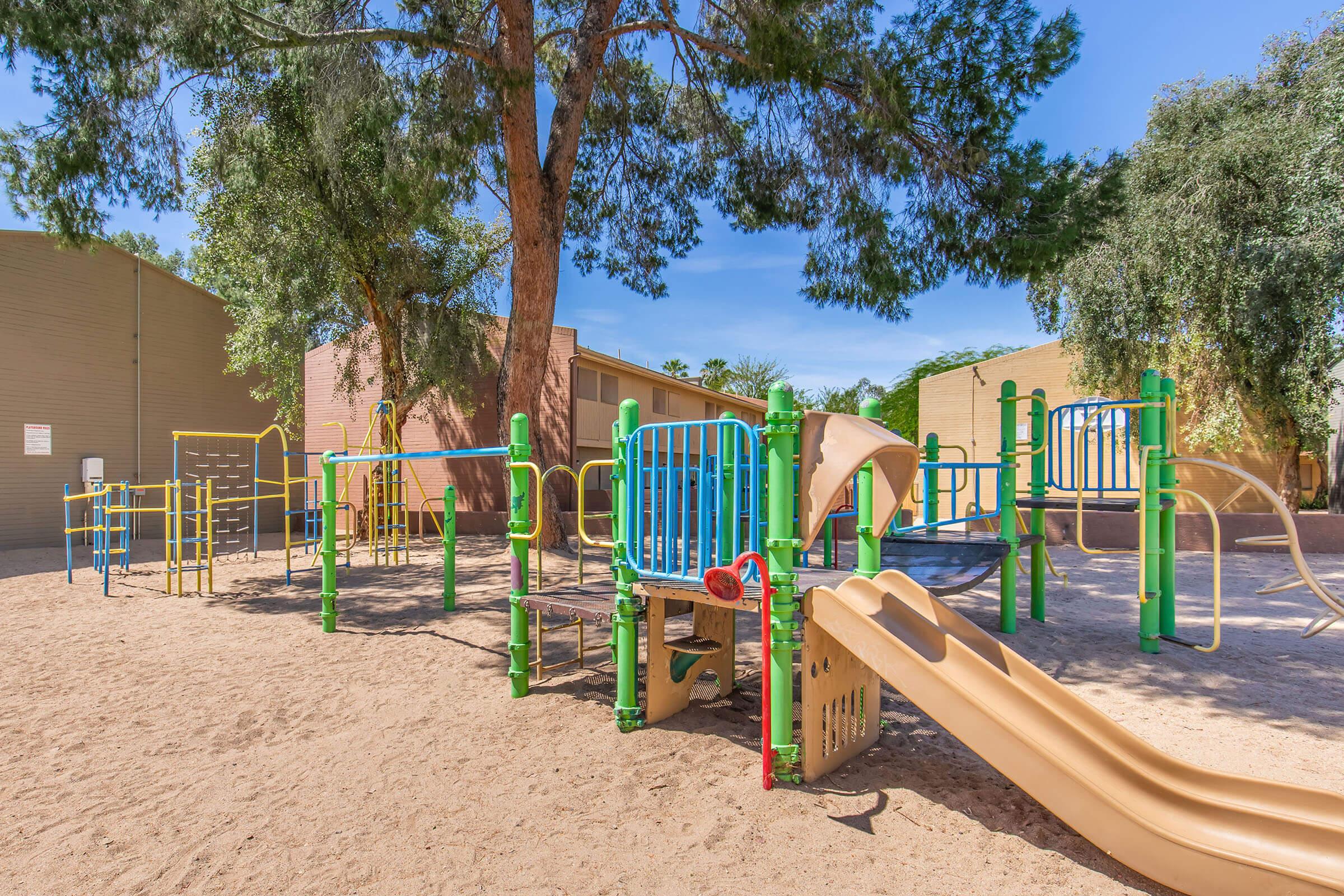 Playground equipment in a sandy area, featuring slides, climbing structures, and colorful railings, under a large tree. Surrounding buildings are visible in the background, with clear blue skies above. The scene is inviting for children to play and explore.