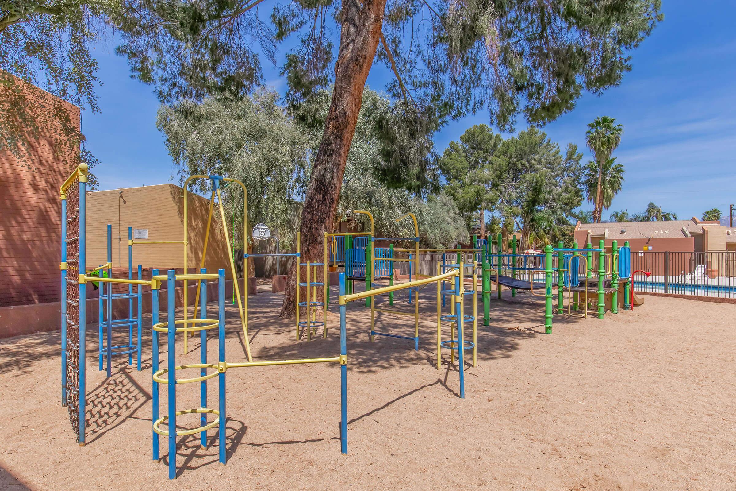 Playground in a sandy area with colorful equipment including climbing structures and slides, surrounded by trees. The background features a building and palm trees, with a clear blue sky overhead.