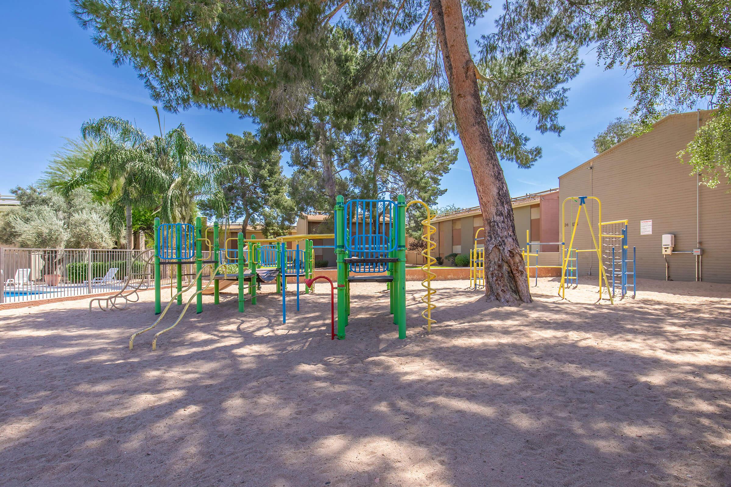 A sunny playground featuring colorful climbing structures and slides, surrounded by sandy ground and trees. The area is fenced with a few chairs nearby, creating a welcoming space for children to play and socialize.