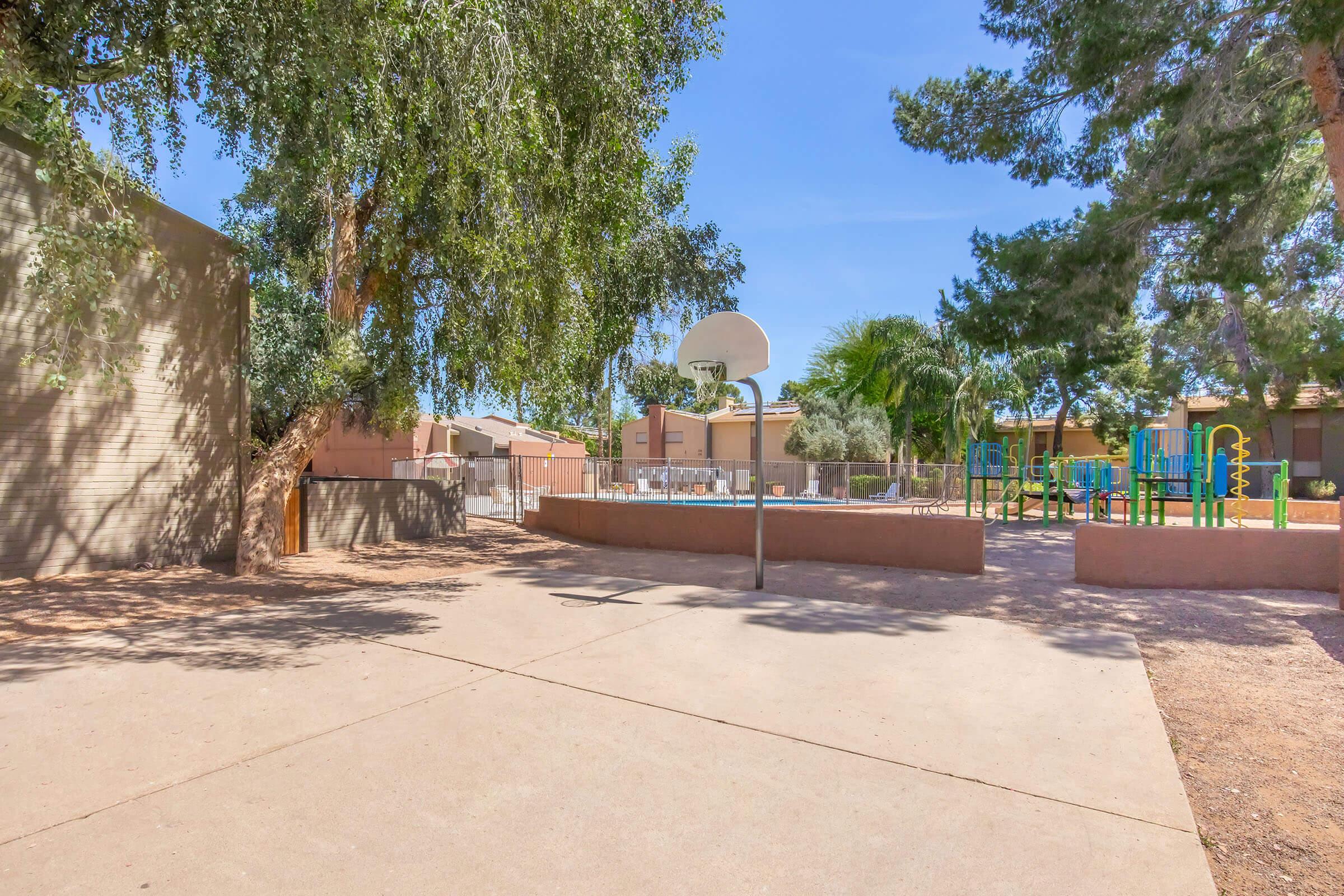 A sunny playground featuring a basketball court surrounded by trees. In the background, there are colorful playground structures and residential buildings. The area is calm and well-maintained, ideal for outdoor activities and recreation.