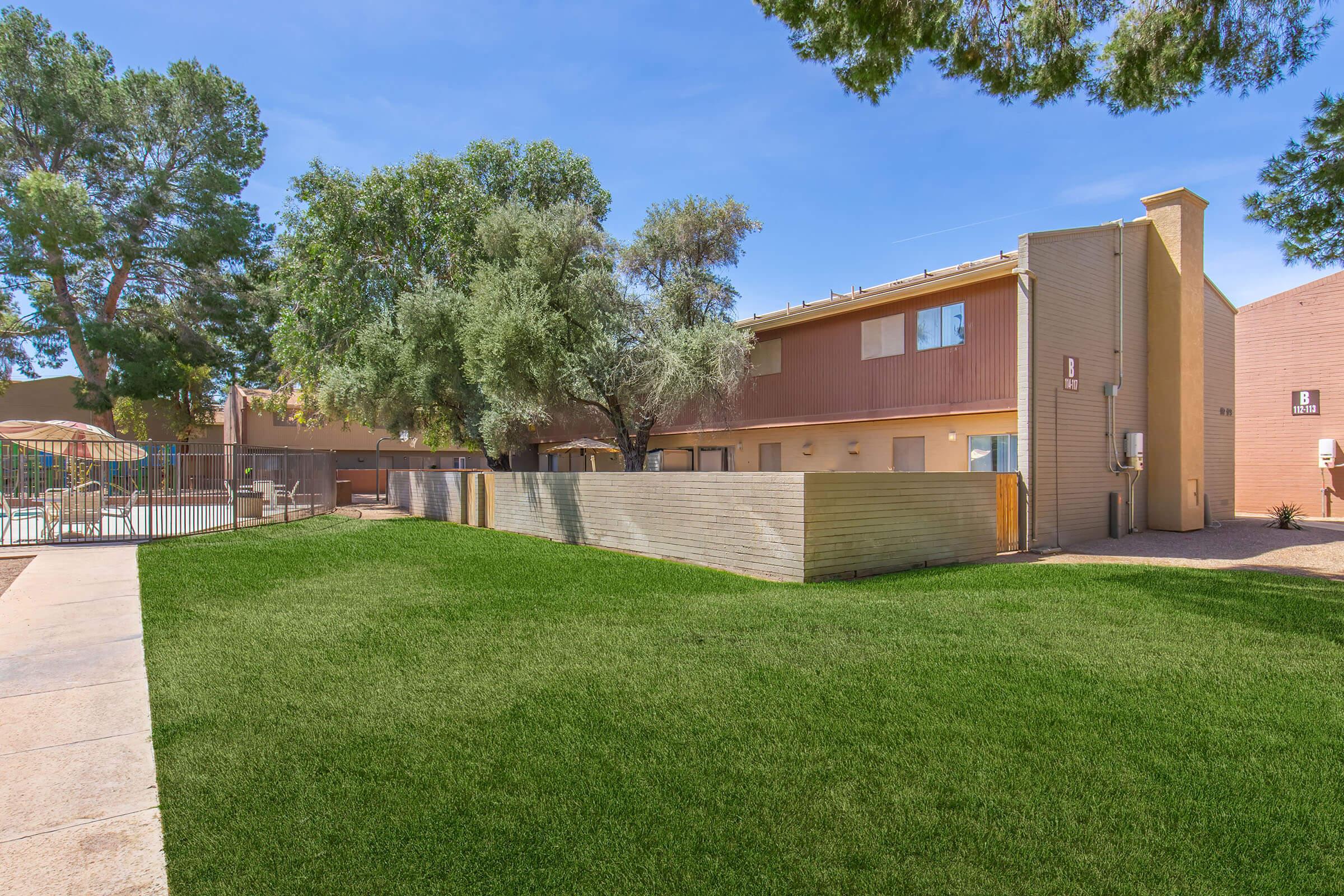 A well-maintained outdoor area featuring a green lawn in front of a two-story residential building. Trees provide shade, and there’s a fenced pool area in the background, indicating a pleasant living environment. Clear blue skies enhance the overall inviting atmosphere.