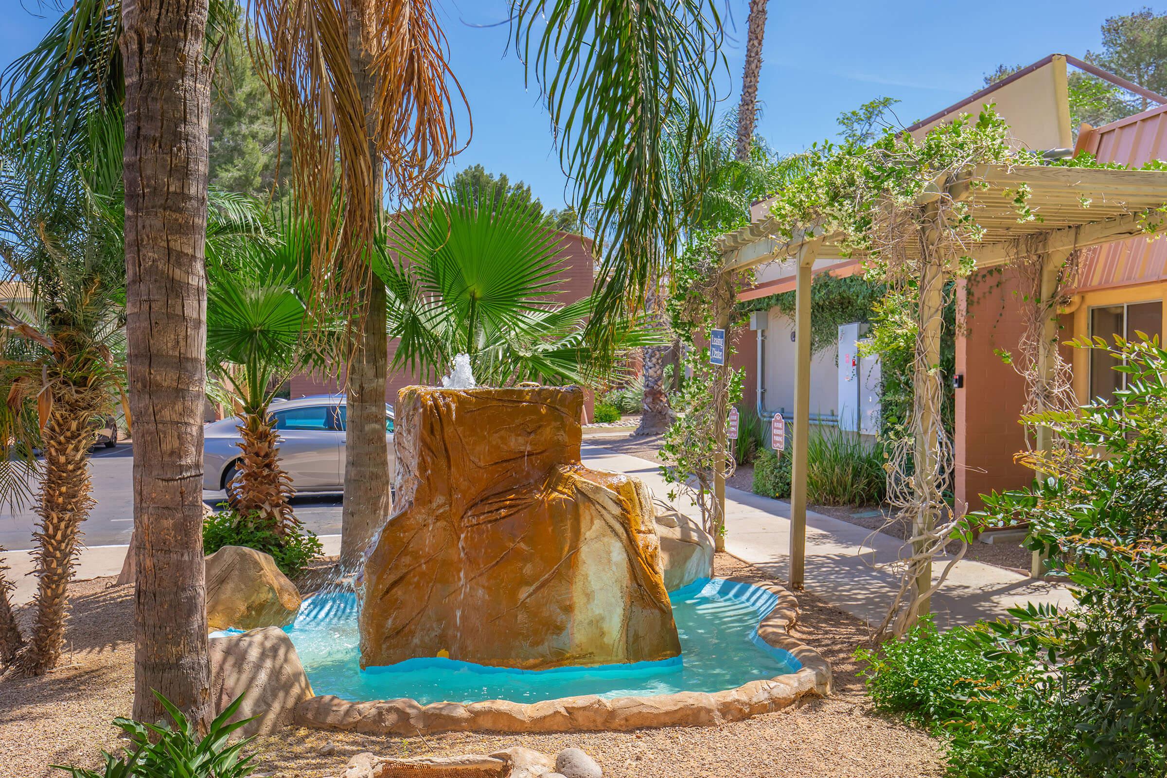 A serene outdoor scene featuring a rock fountain surrounded by palm trees and lush greenery. The water cascades gently from the top of the rocky structure into a turquoise pool at the base. Nearby, a pathway leads to a building, and sunlight filters through the leaves, creating a peaceful ambiance.