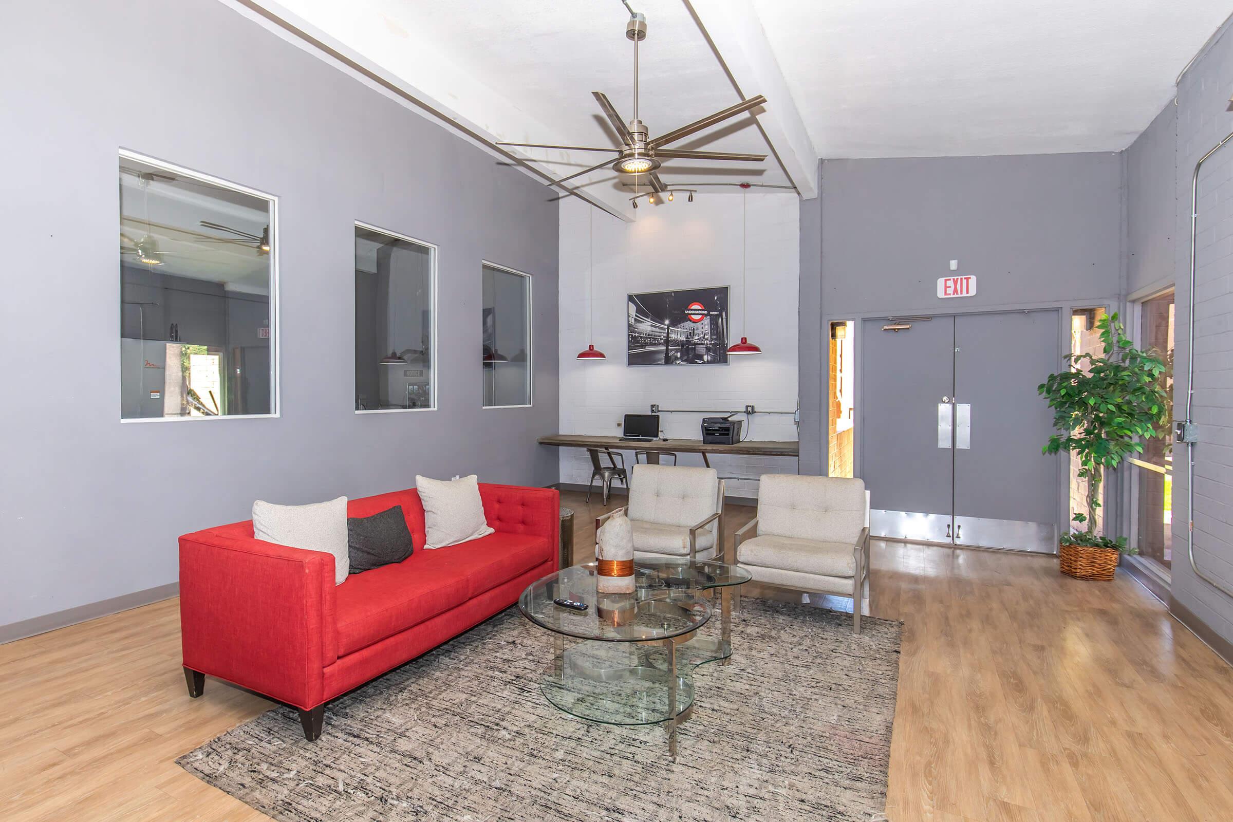 A modern living room featuring a bright red sofa with gray and white accent pillows, a glass coffee table, and two light-colored chairs. The walls are painted gray with large windows. A ceiling fan hangs overhead, and there's a minimalist desk area in the background with a photograph on the wall.
