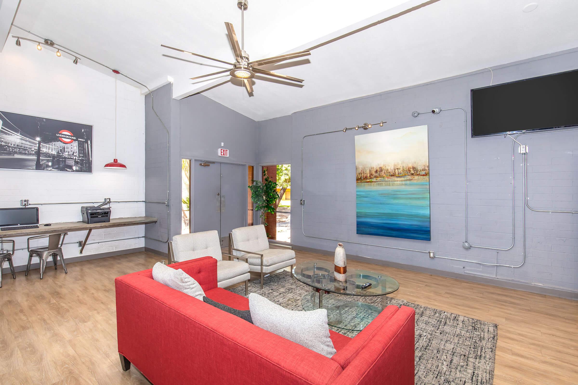 Modern living room with a red sofa and gray armchairs around a glass coffee table. The walls are painted gray, featuring an abstract blue painting and a large TV. A ceiling fan hangs above, and there is a workspace with metal stools and a printer in the corner. Bright lighting and wood flooring enhance the inviting atmosphere.