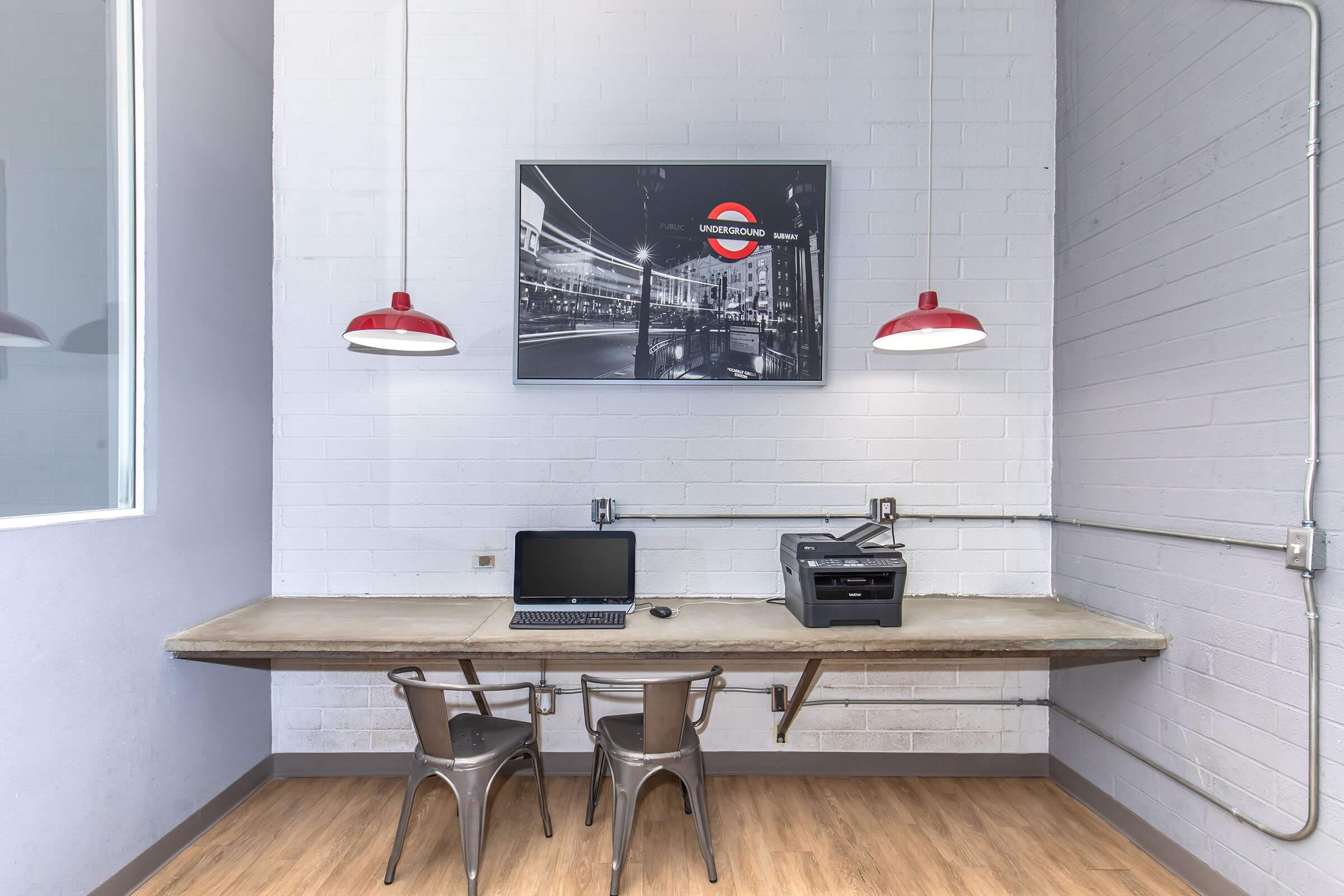 A modern workspace featuring a large table with a laptop and a printer. Two metallic chairs are positioned at the table. Above, a black-and-white photograph of an underground train station is displayed on the wall, flanked by two red pendant lights. The walls are painted in light gray, enhancing the contemporary feel of the room.