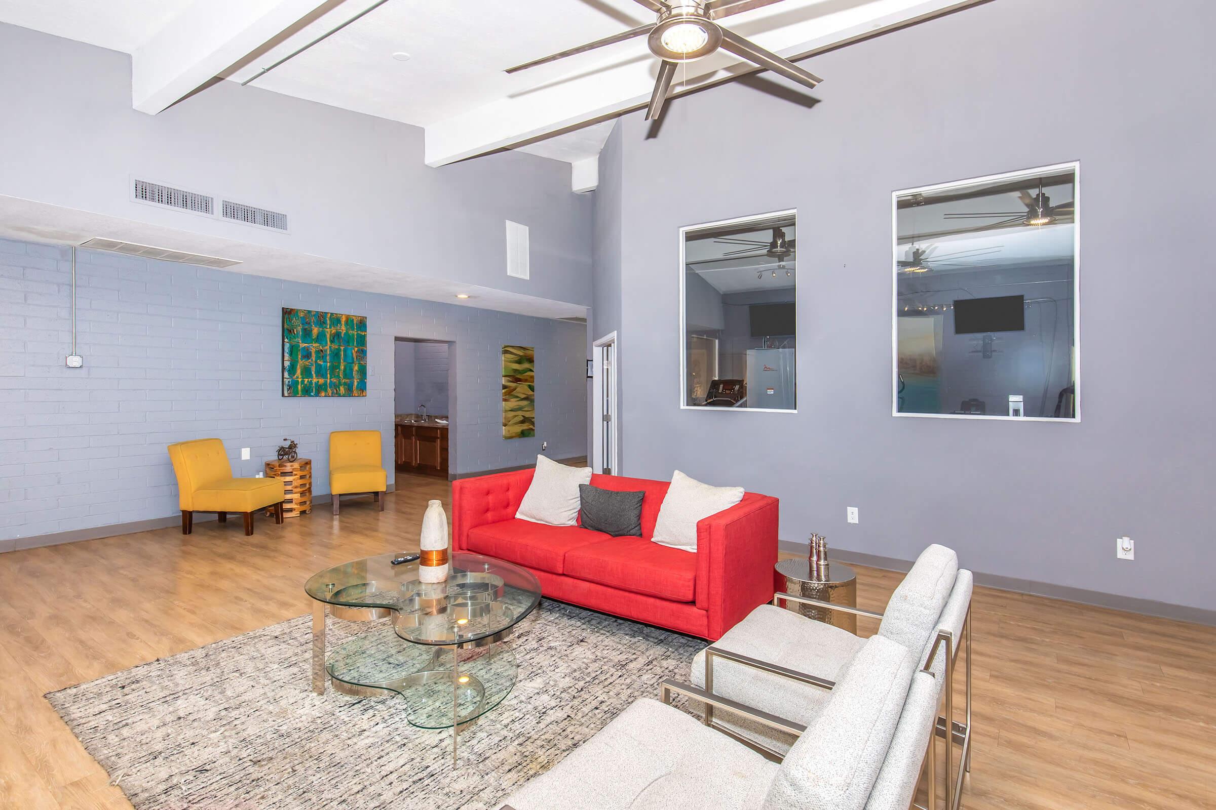 A modern living room featuring a red sofa with cushions, gray armchairs, a glass coffee table, and a large area rug. The walls are painted in light gray, and there are two large mirrors and ceiling fans. Additional seating is visible in the background, along with artwork on the walls.
