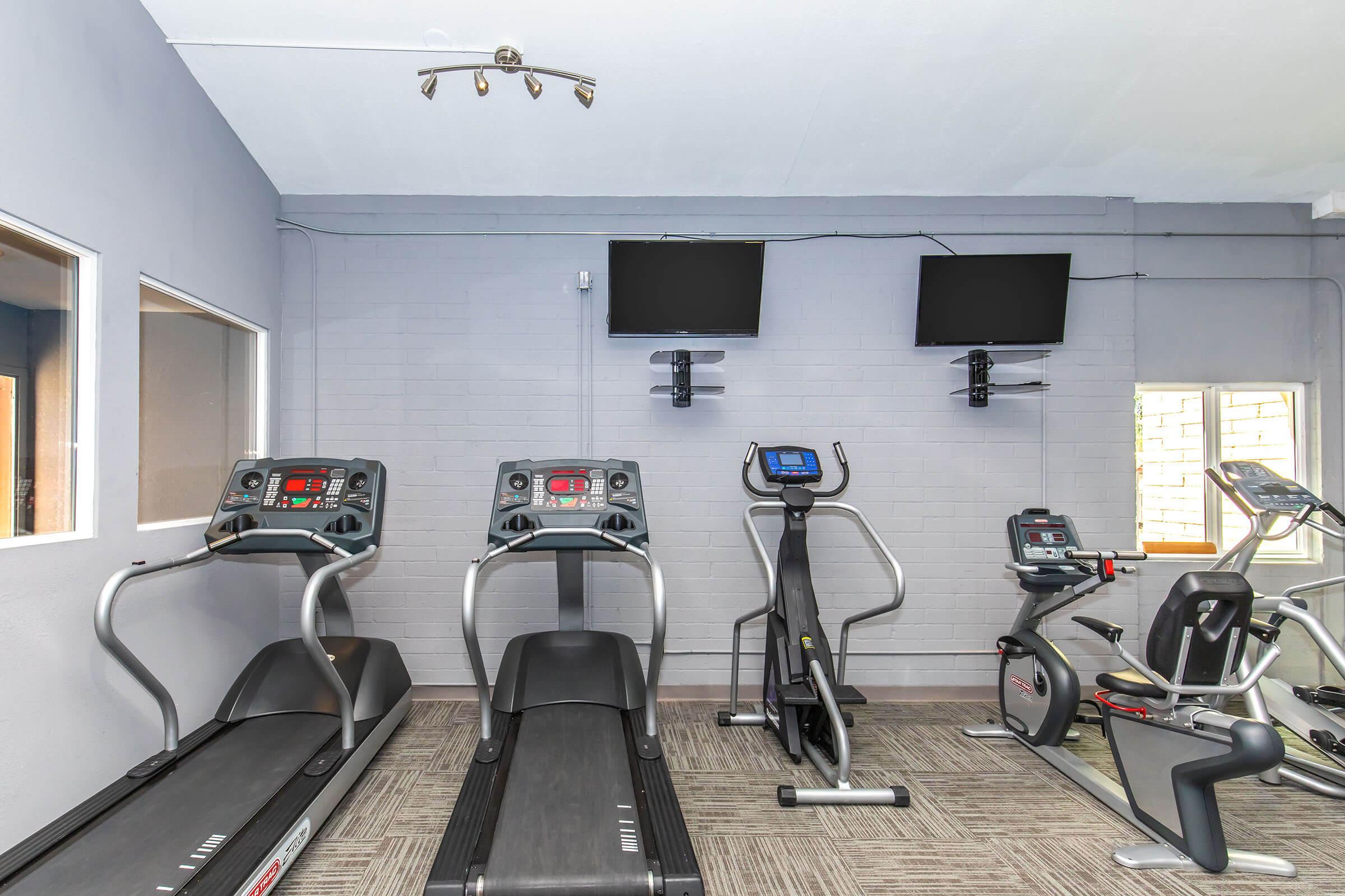 A modern gym area featuring two treadmills, an elliptical machine, and a stationary bike. The walls are painted gray, and there are two flat-screen TVs mounted above the equipment. Natural light enters through windows on the side, creating a bright, inviting space for exercise.