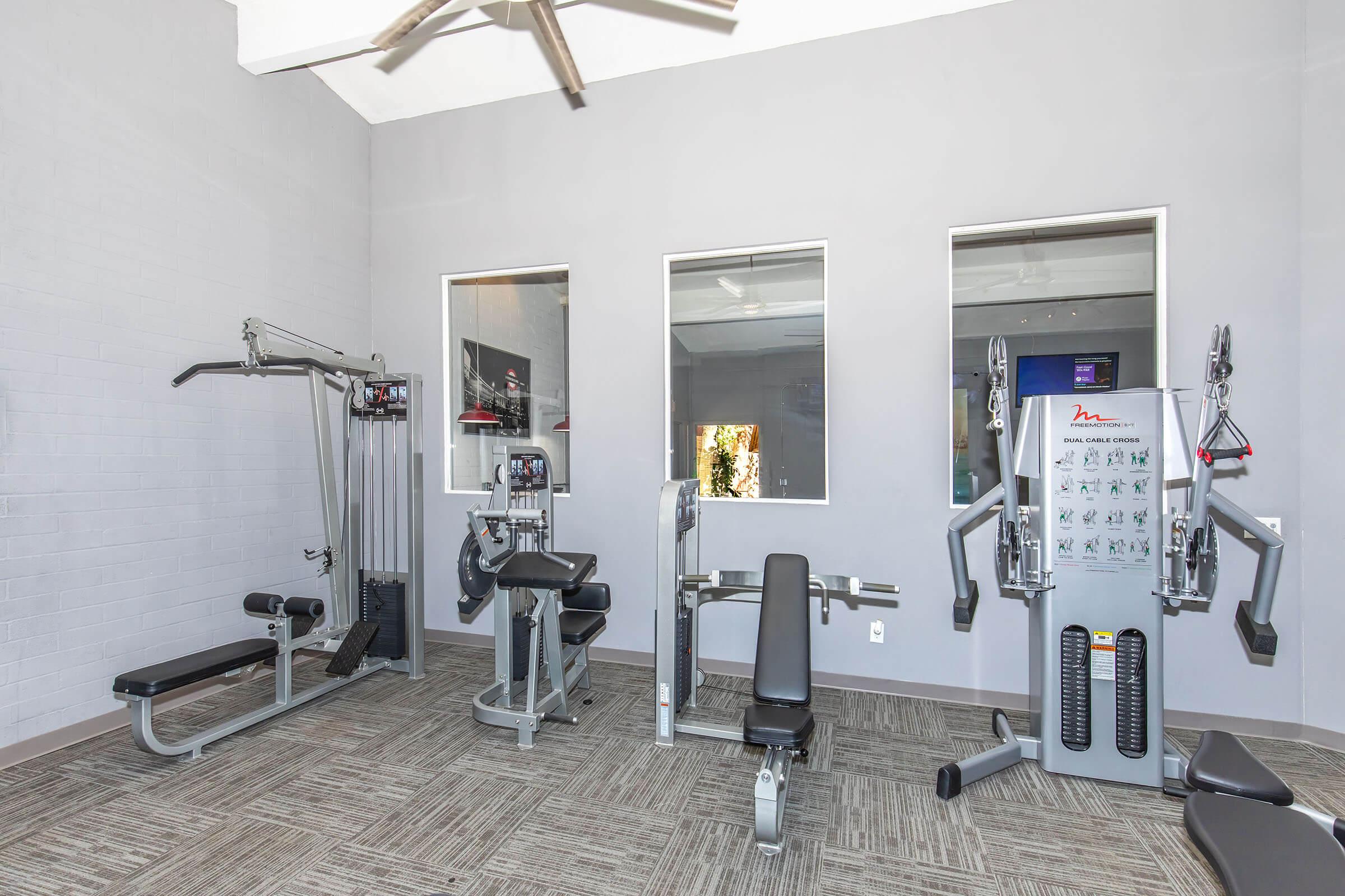 A modern gym interior featuring various exercise equipment, including a multi-station machine, a bench, and weights. The walls are painted in a neutral color, and large windows allow natural light to fill the space. The floor is carpeted with a textured pattern, creating a comfortable workout environment.