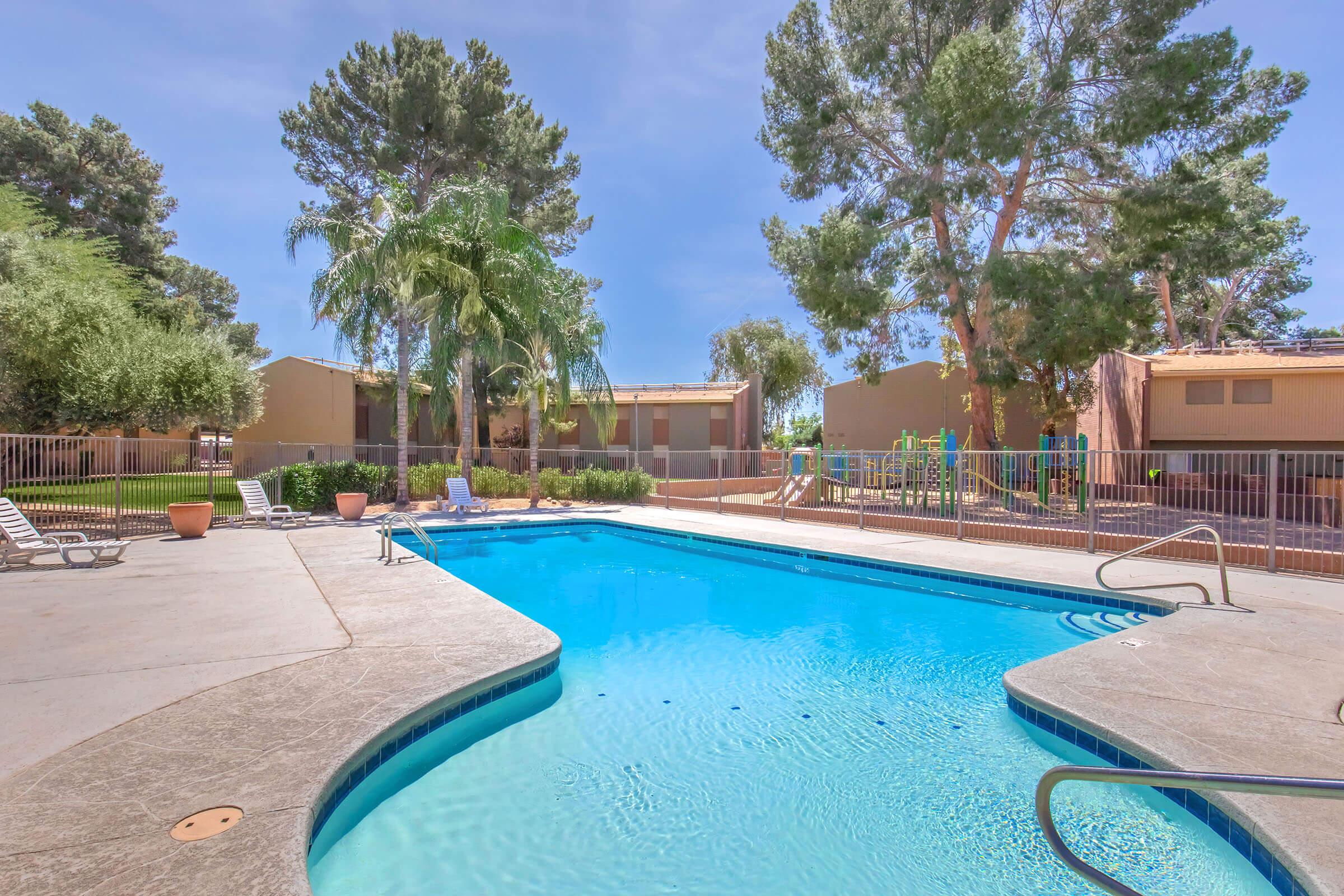 A clear, inviting swimming pool surrounded by palm trees and lounge chairs. In the background, there are residential buildings and a playground. The sky is sunny, creating a bright and relaxing atmosphere.