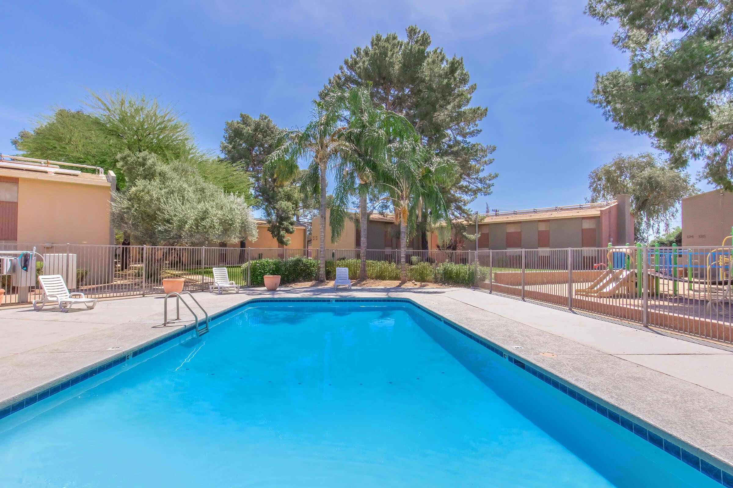 A clear blue swimming pool surrounded by a fenced area, with lounge chairs on the pool deck. Lush green trees and shrubs are visible in the background, alongside buildings with closed shutters. The scene is bright and sunny, typical of a residential or recreational area.