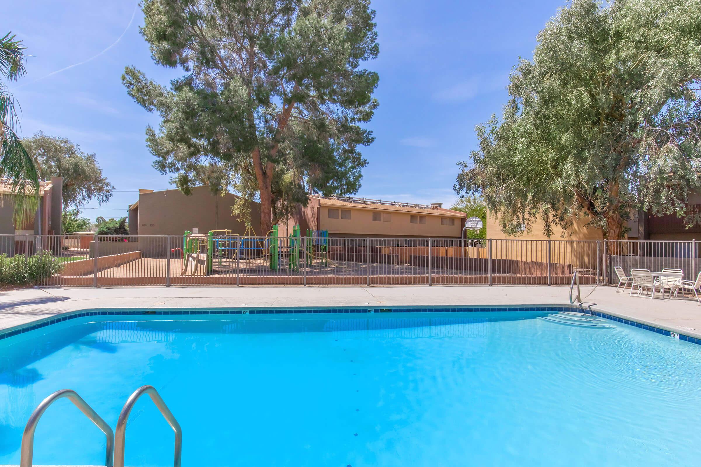 A clean swimming pool surrounded by a fence, with trees and a playground in the background. The area features beige buildings and blue skies, creating a tranquil outdoor setting. The pool area is well-maintained, inviting relaxation and leisure.