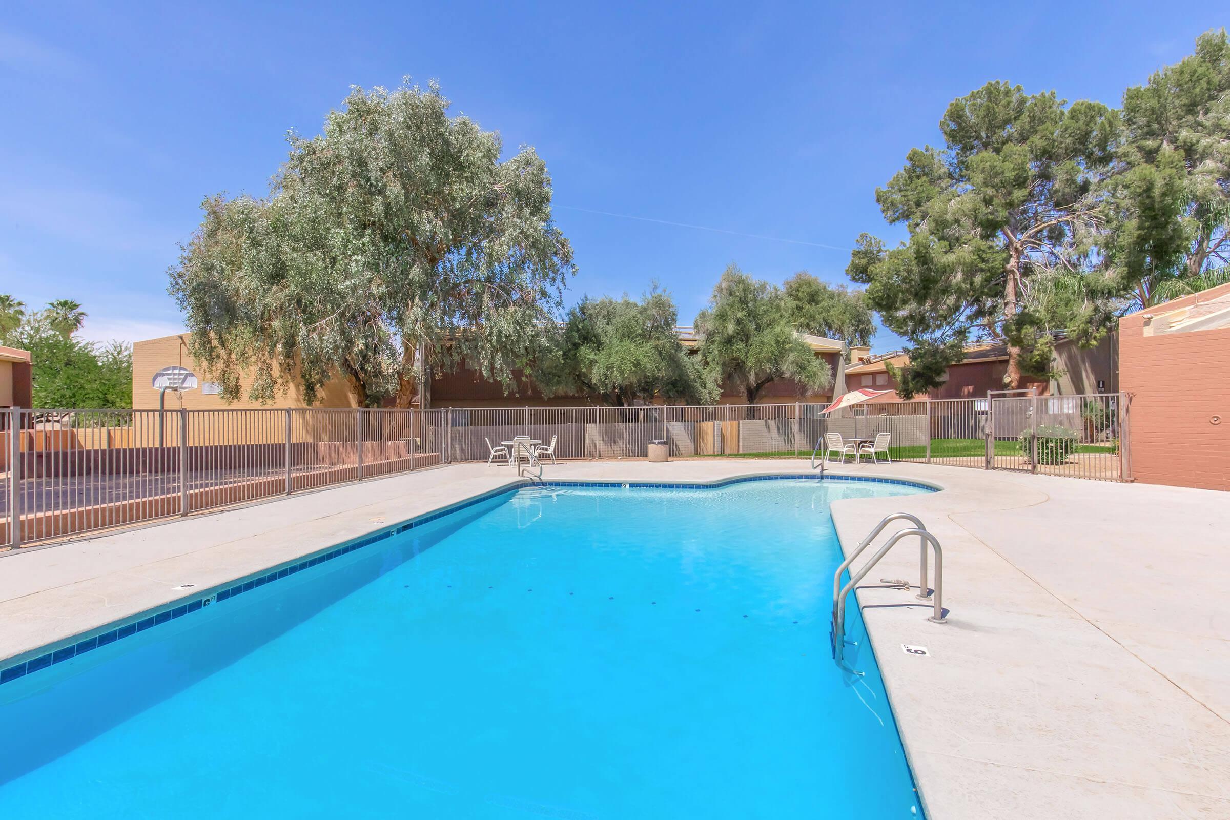 A clear swimming pool surrounded by a concrete deck, with a few lounge chairs on the side. Lush green trees provide shade near the pool area, and a fence encloses the space. In the background, there are buildings and more greenery under a blue sky.