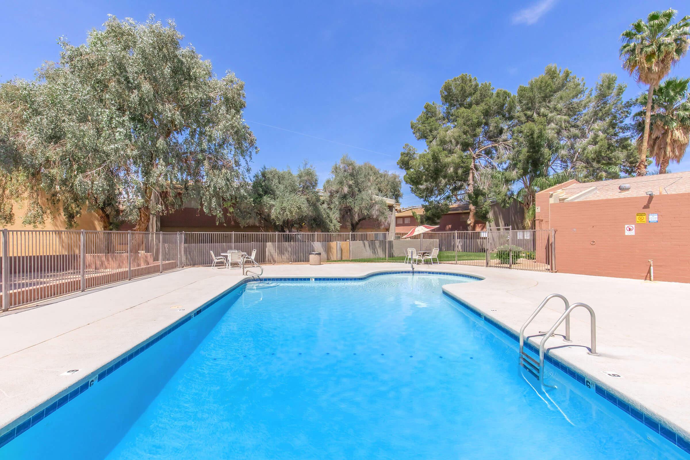 A sparkling blue swimming pool with a diving board, surrounded by a light-colored deck. There are a few lounge chairs placed around the pool area. Lush trees and shrubbery provide shade in the background, while a clear blue sky overhead enhances the inviting atmosphere of the outdoor space.