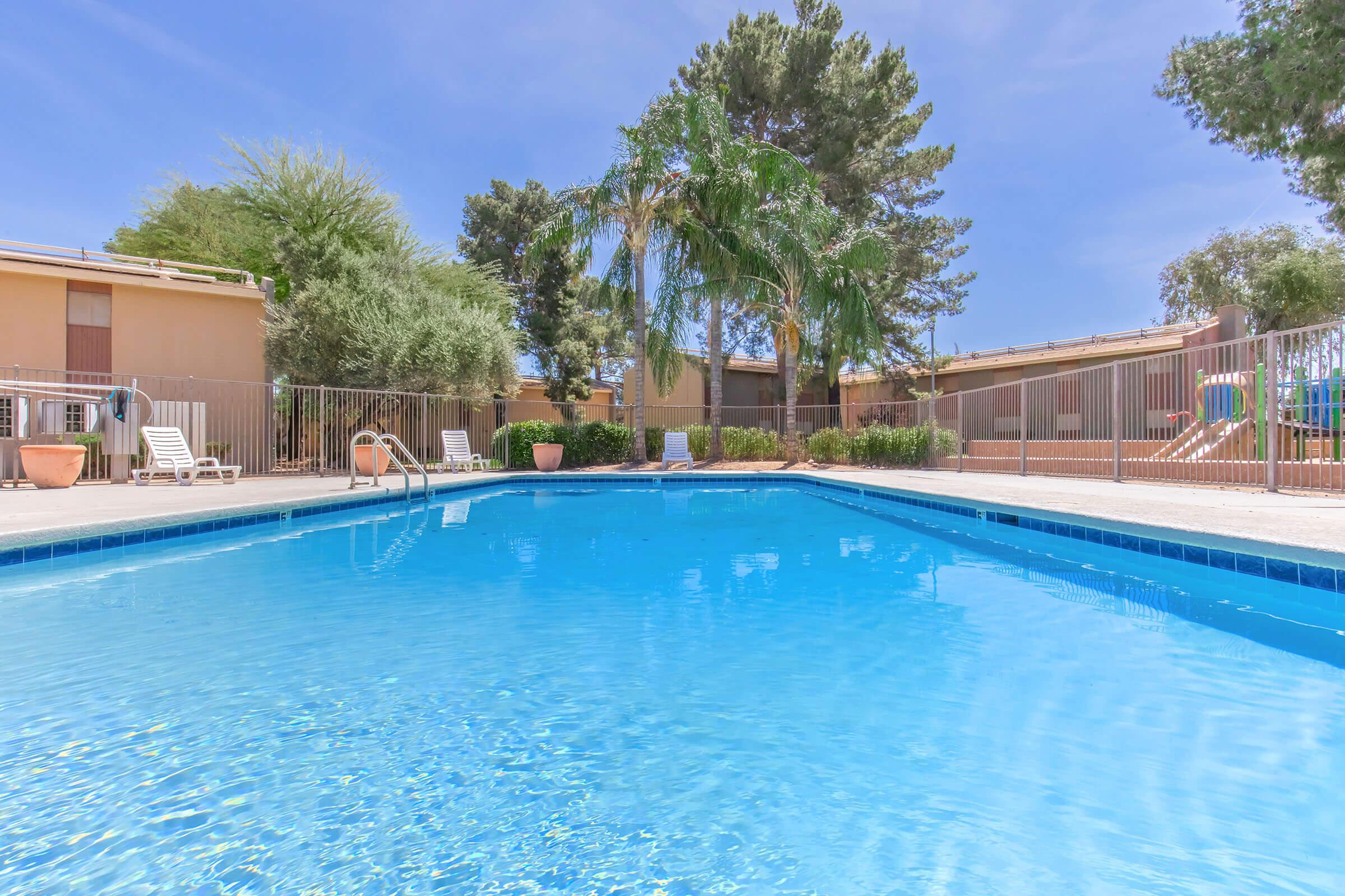 A clear blue swimming pool surrounded by lounge chairs and palm trees, with a sunny sky in the background. The pool area features a fence, and nearby are buildings with a warm color tone, creating a relaxing outdoor atmosphere.
