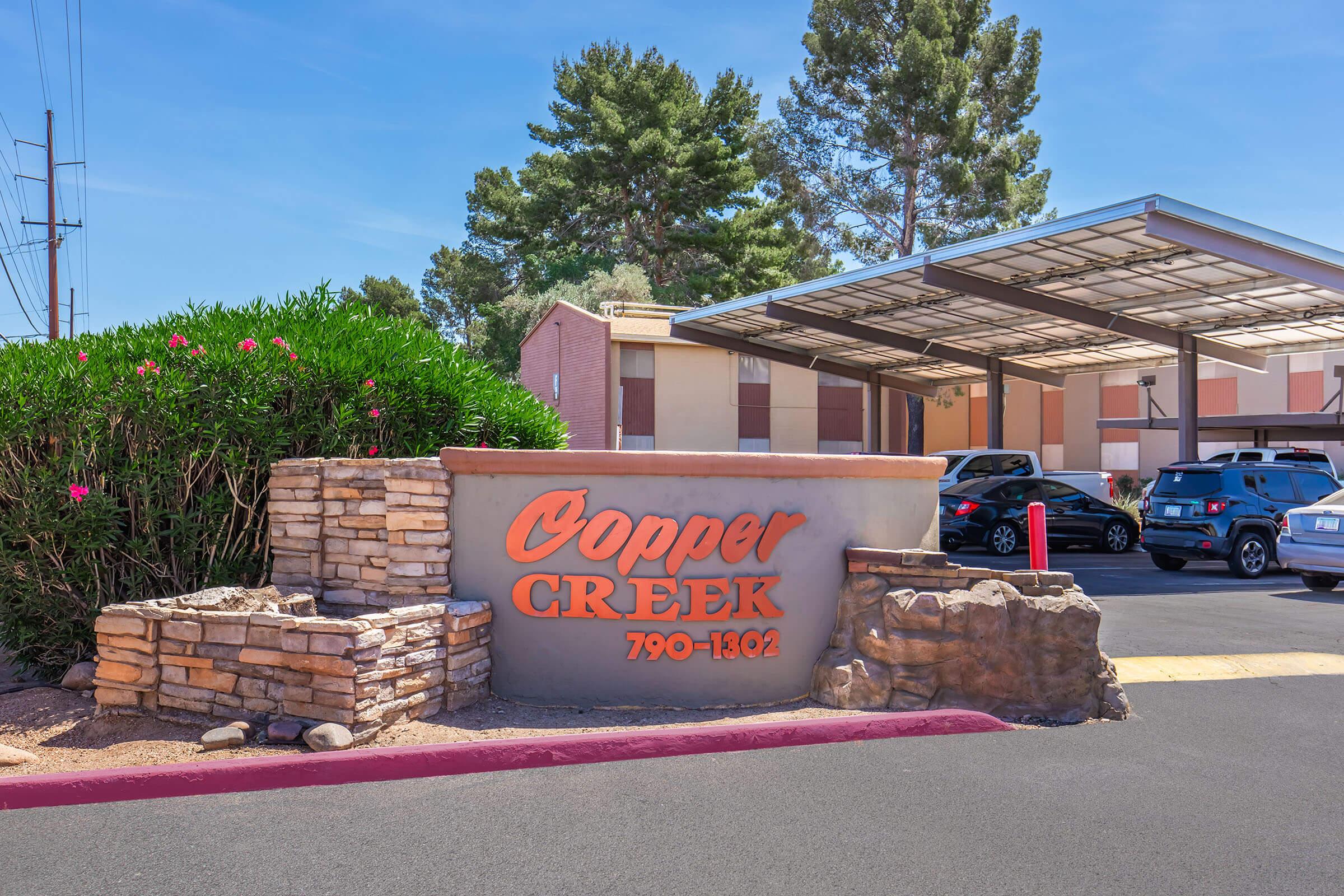 Sign for Copper Creek featuring bold orange lettering on a stone base, surrounded by greenery. The background shows a multi-panel building with a carport and parked cars under a clear blue sky. The address listed is 790-1802.