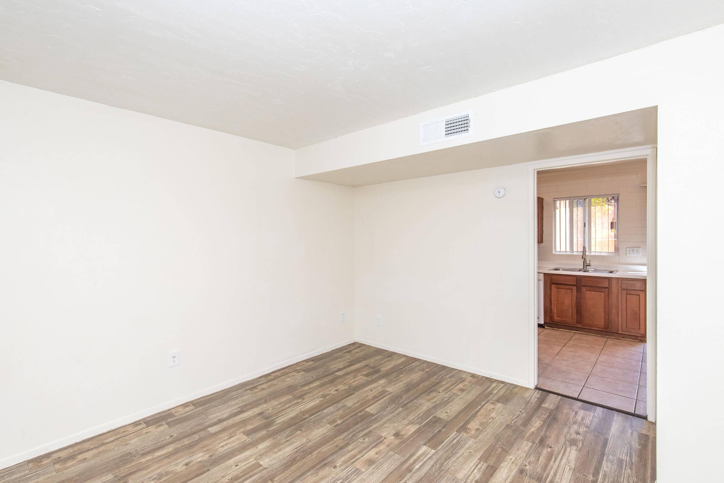 A spacious, empty room with light-colored walls and a wood-like floor. A doorway on the right leads to a kitchen area with brown cabinets. Natural light enters through a window in the kitchen, creating a bright and open atmosphere. The overall design is simple and modern, ideal for various decoration styles.