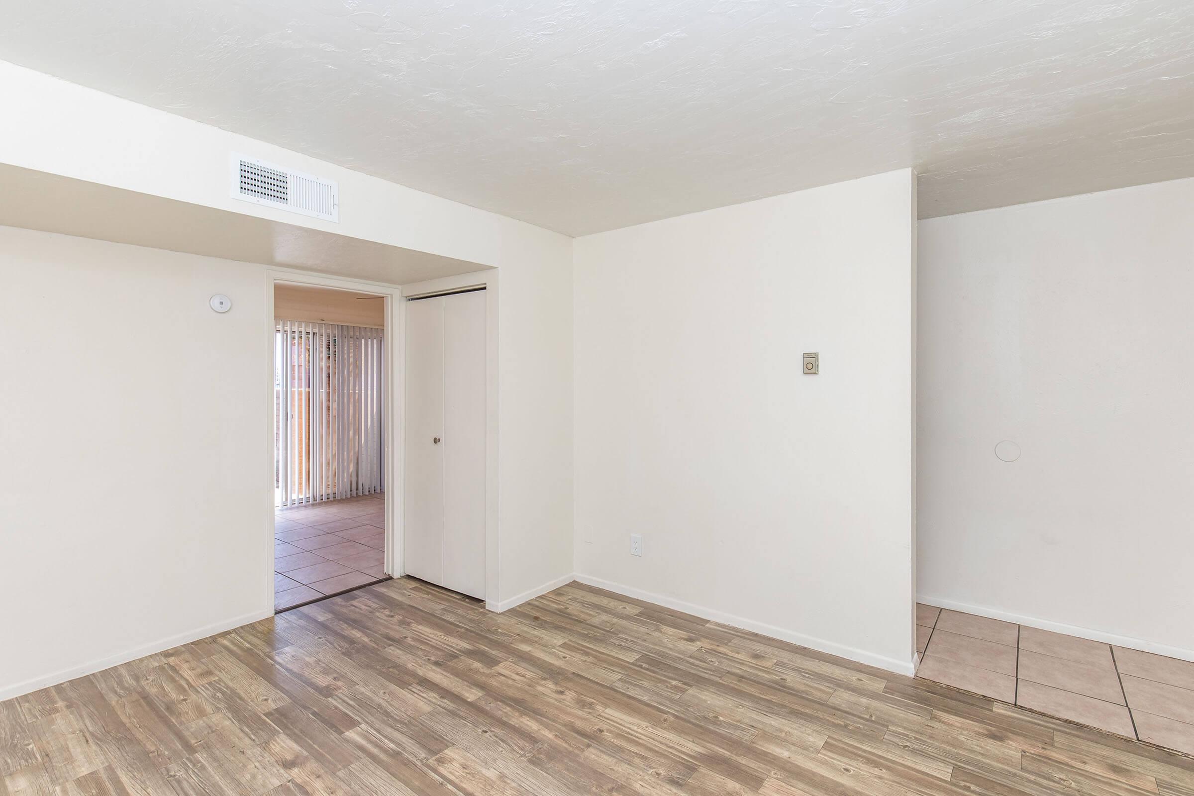 A bright, empty interior space showcasing light-colored walls and a wood-like floor. A doorway leads to an area with sliding glass doors and beige tile flooring. The room features a small closet and is well-lit, suggesting a welcoming and open atmosphere.