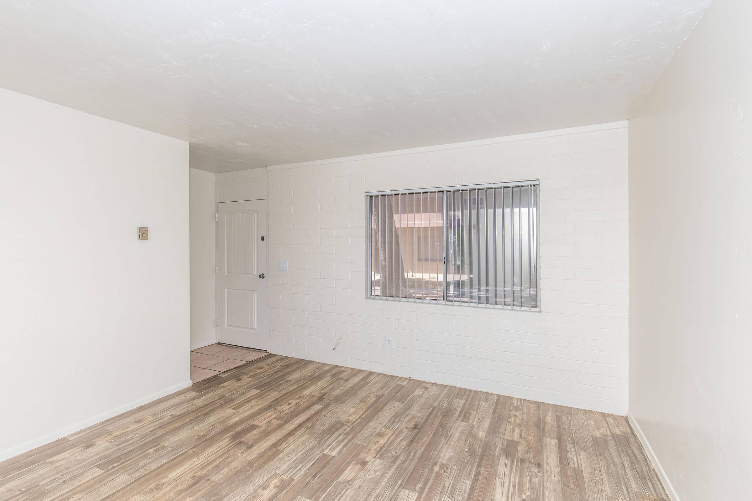 A spacious, empty room with light-colored walls and wooden flooring. There's a large window with blinds letting in natural light, and a door leading to another area. The setting appears clean and minimalistic, suitable for various uses like a living room or office space.