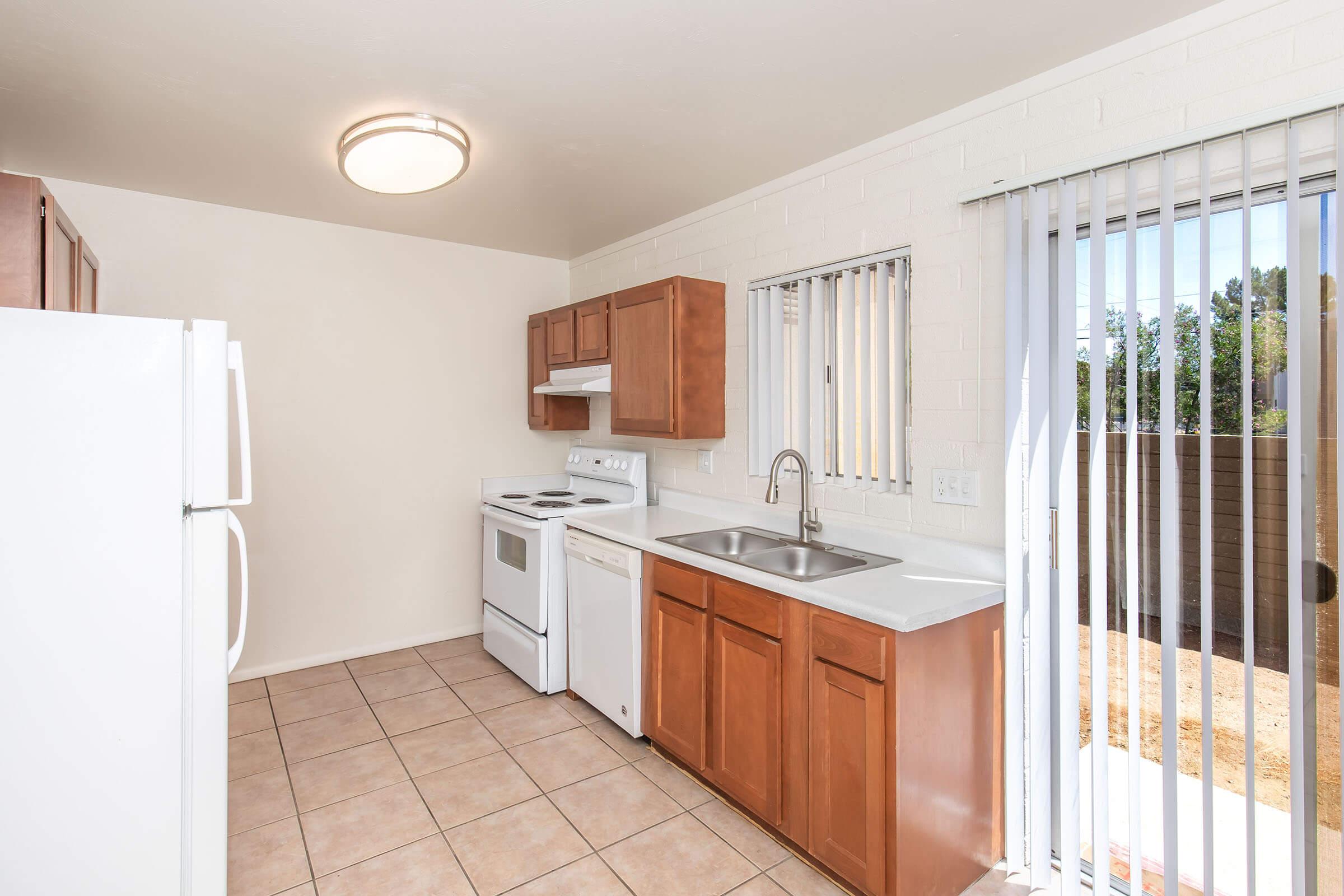 A small kitchen featuring wooden cabinets, a white refrigerator, an oven, and a sink with a countertop. There are blinds on a window and a door leading to an outdoor area. The tiled floor enhances the space, creating a clean and functional environment for cooking.