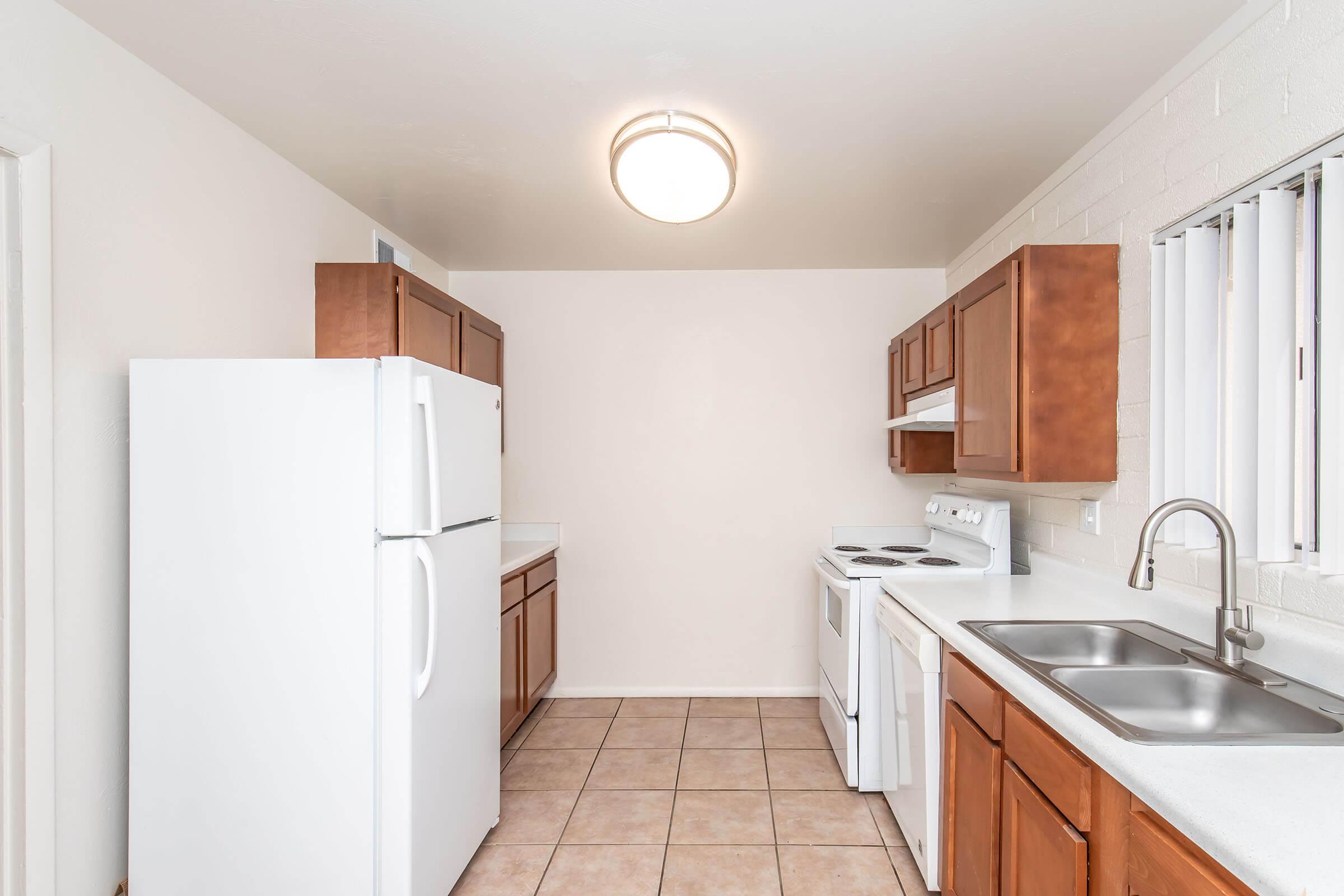 Bright kitchen with light-colored walls and cabinetry. Features include a white refrigerator, stove, and sink with a long countertop. The floor is tiled, and there is a window with blinds allowing natural light. A modern light fixture hangs from the ceiling, adding brightness to the space.