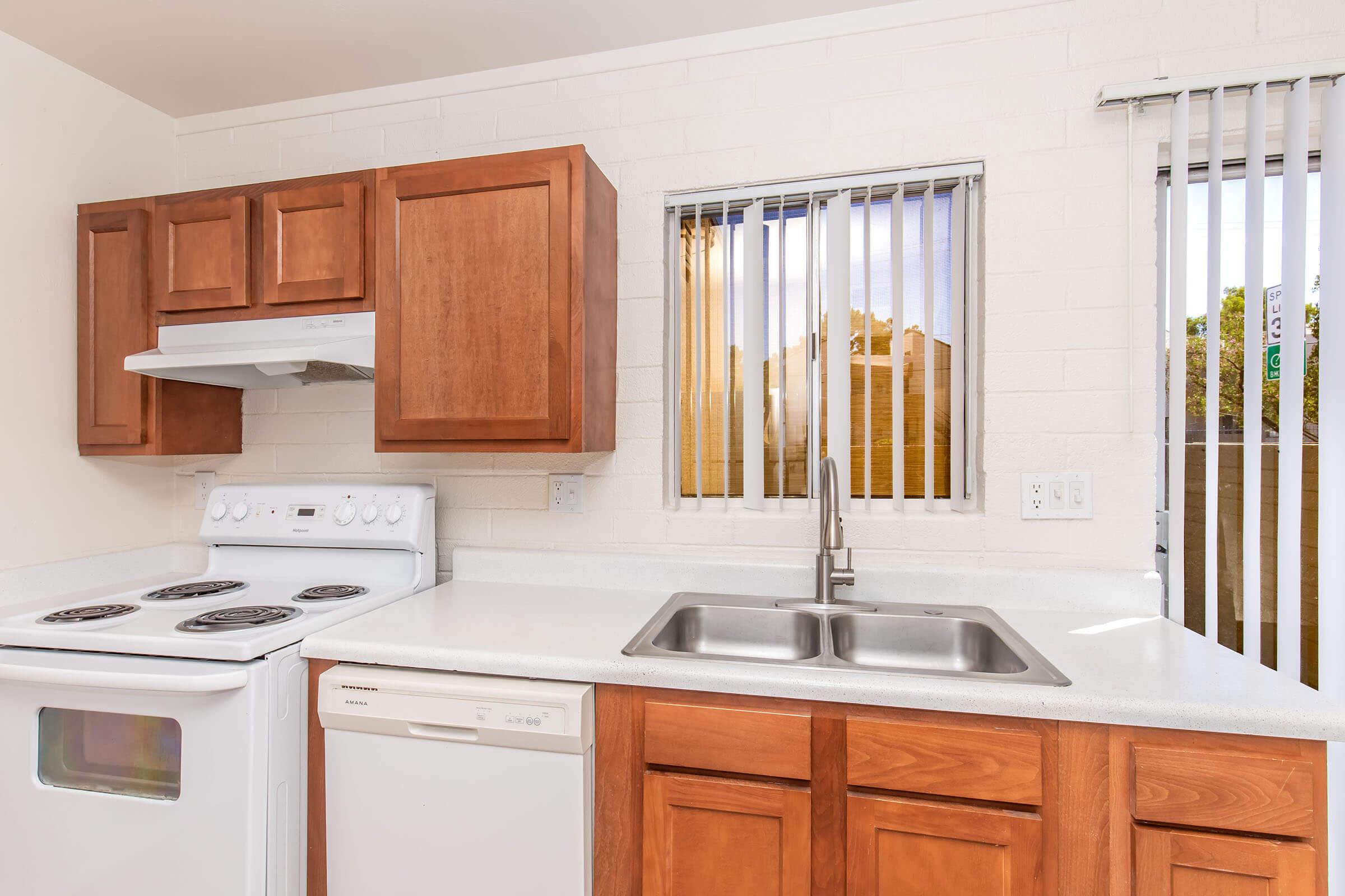 A modern kitchen featuring brown wooden cabinets, a white countertop, and stainless steel sink. It includes an electric stove, a dishwasher, and two windows with blinds, allowing natural light to illuminate the space. The overall design is bright and functional.