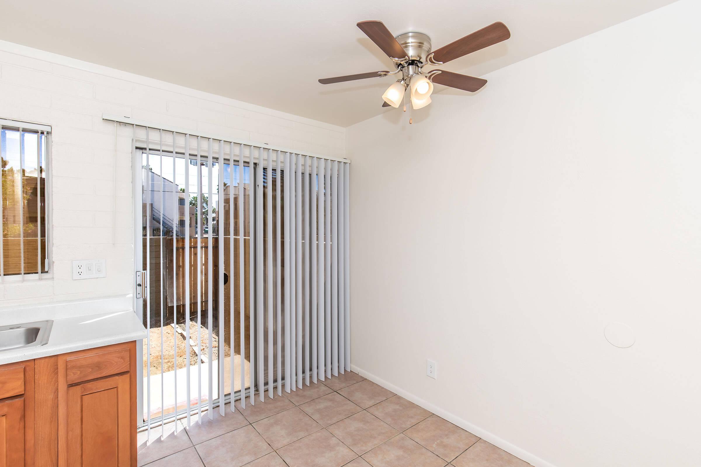 A small, bright room with tiled flooring, featuring a ceiling fan with three lights. On one side, there is a window with vertical blinds, and a sliding glass door leads to a fenced outdoor area. The walls are painted white, and there's a compact kitchen sink with wooden cabinetry.