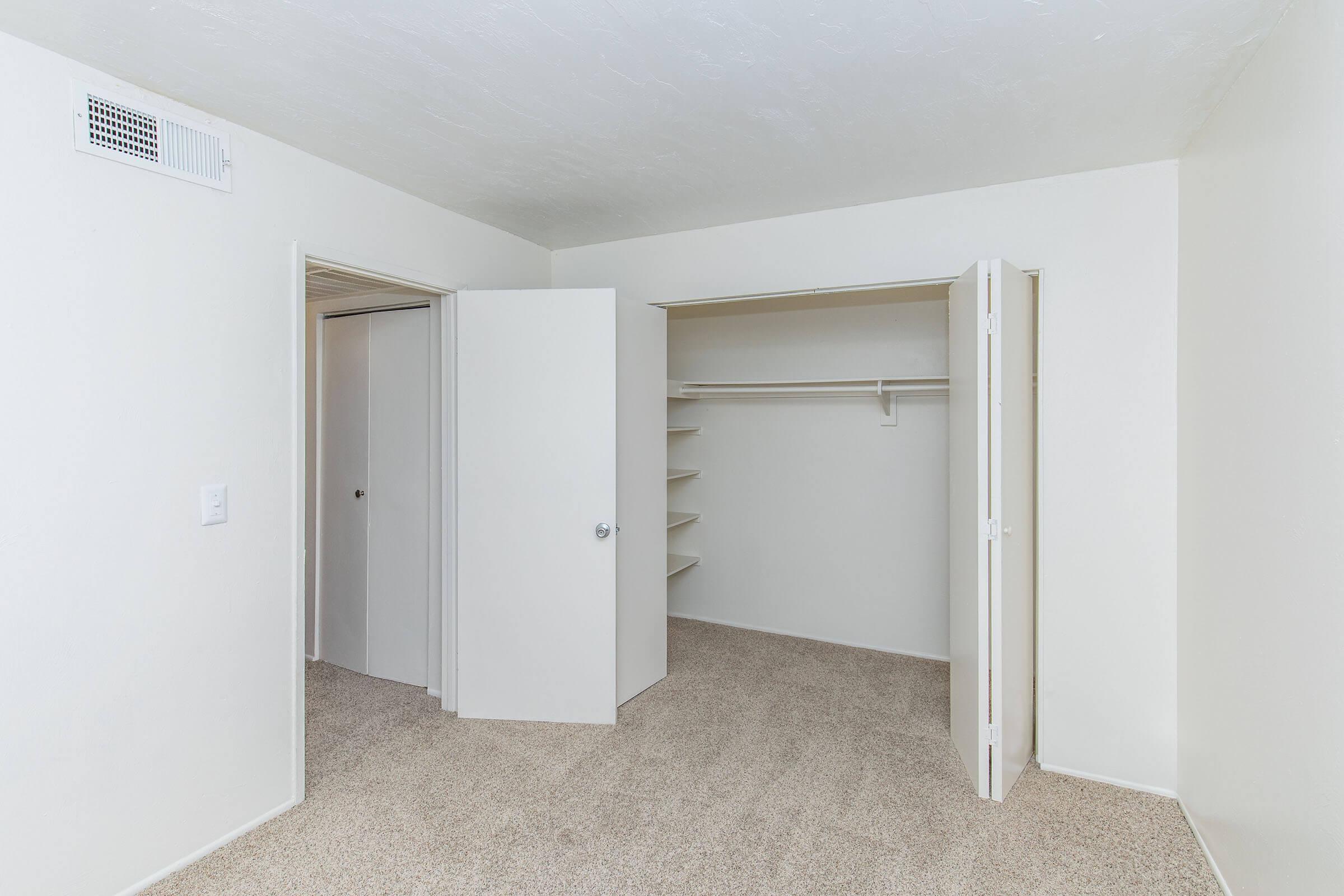 A spacious empty room with beige carpeting and light-colored walls. There is a closet with double doors on the left side, featuring shelves and a hanging rod. A door leading to another room is visible on the right. Natural light fills the space, creating a bright and clean atmosphere.