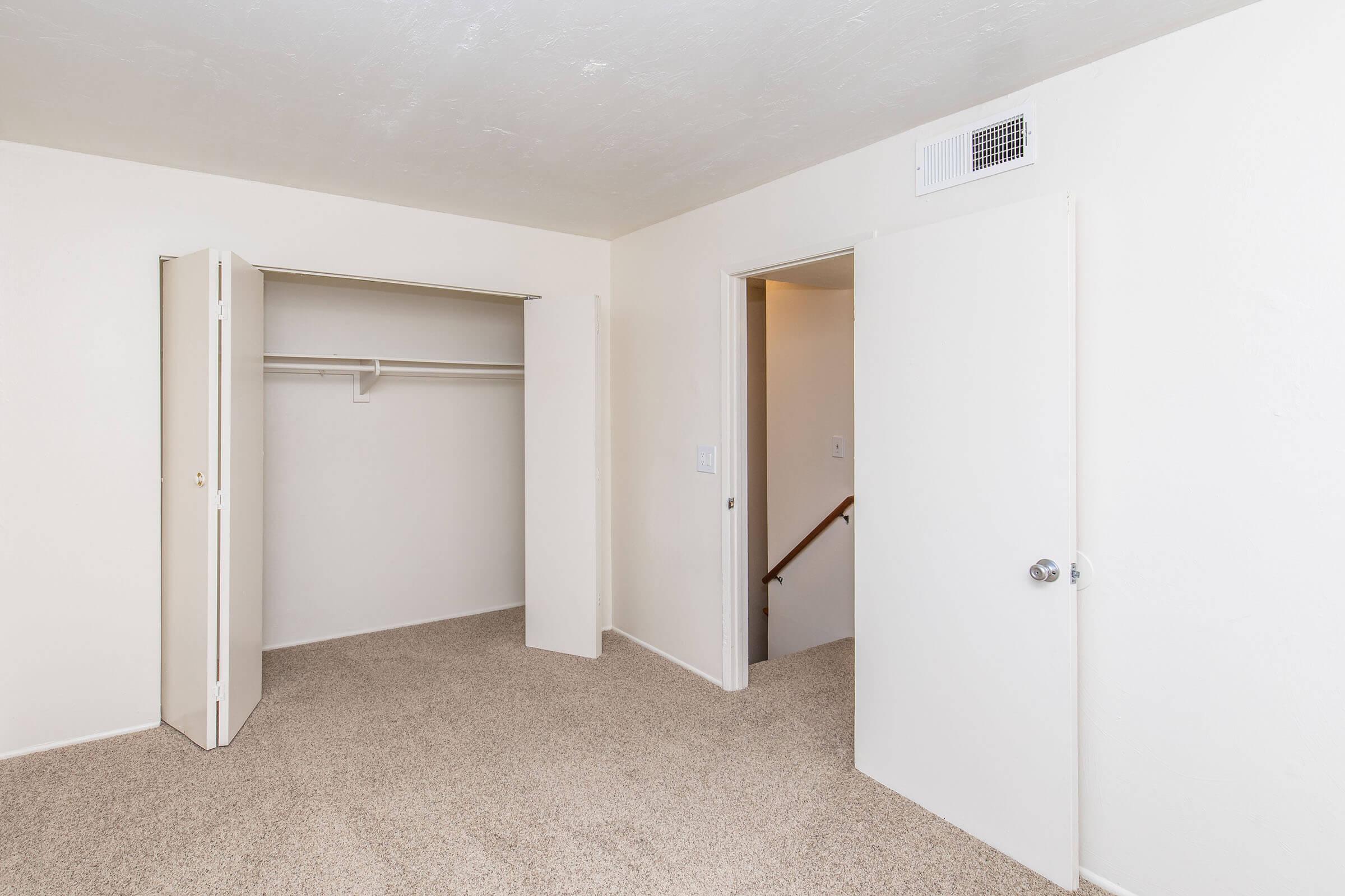 A light-colored, empty room featuring beige carpet flooring. There is an open closet with sliding doors on one side and a closed white door leading to another area on the opposite side. The walls are painted in a neutral tone, and a small vent is visible near the ceiling.