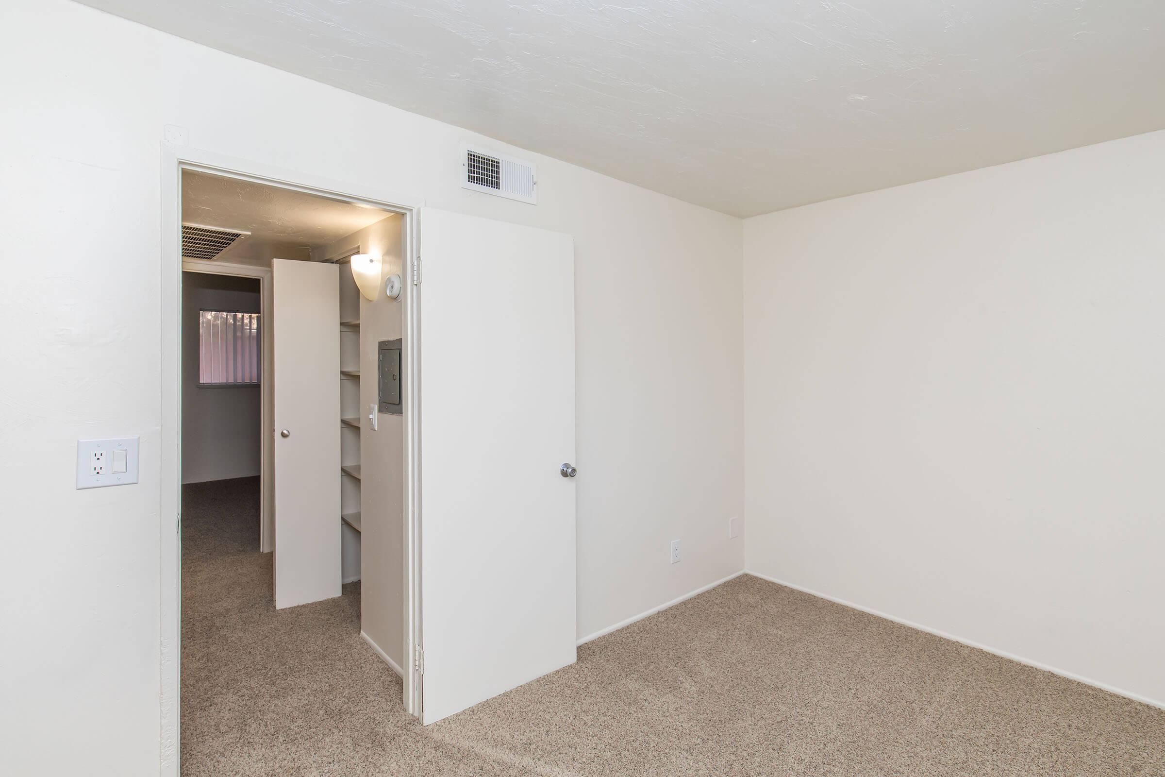 A simple, empty room with beige carpet and light-colored walls. The room features an open door leading to a small hallway and another room, and a built-in shelf on the wall. Natural light enters through the hallway, creating a bright and airy atmosphere.