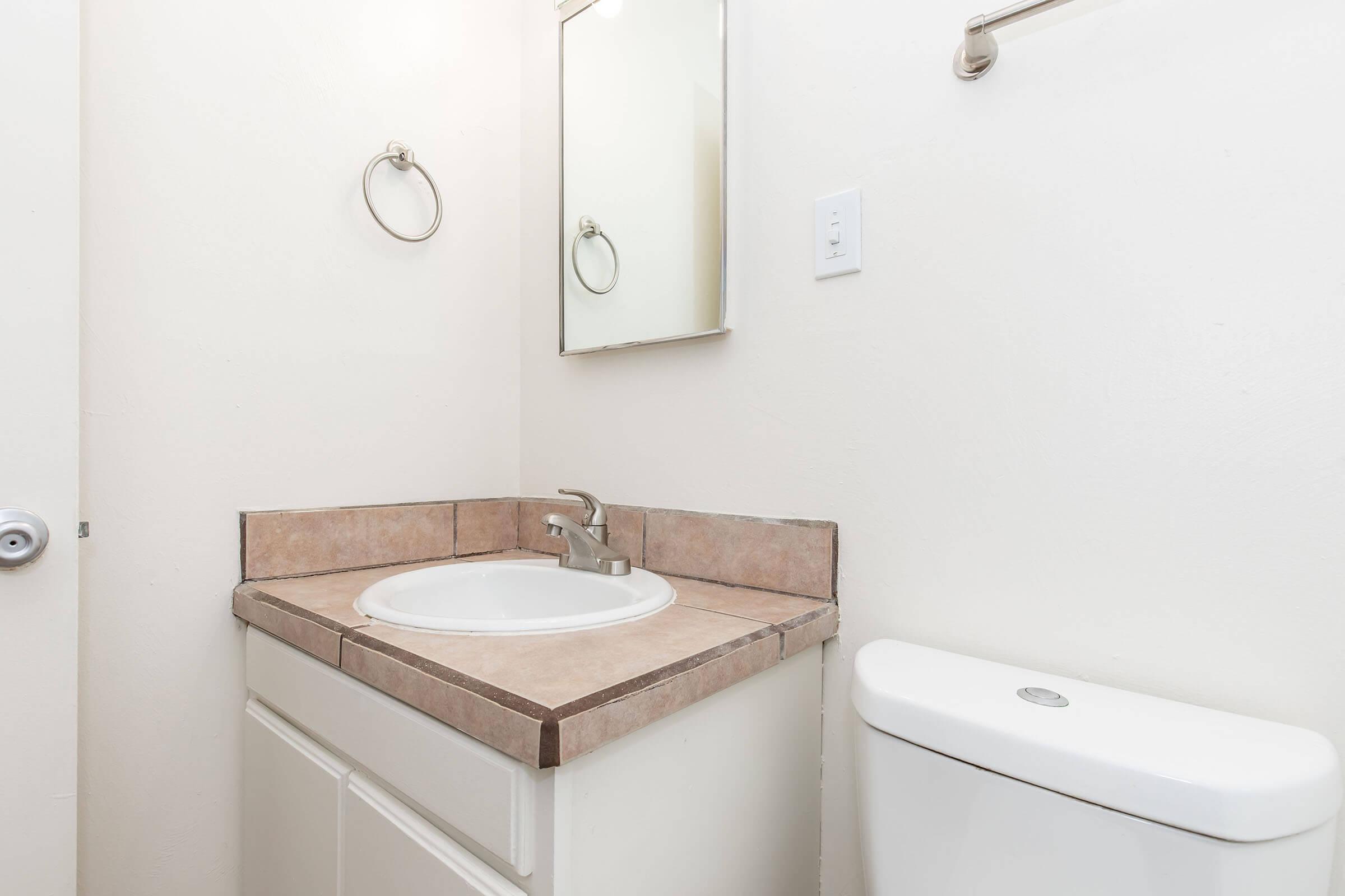 A small bathroom featuring a sink with a circular basin and faucet on a tiled counter, a mirror above the sink, a toilet nearby, and a towel rack. The walls are painted in a light color, creating a clean and simple aesthetic.